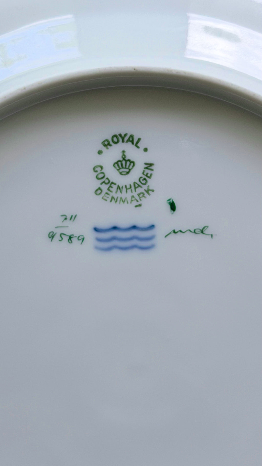 The underside of a white porcelain plate or dish showing Royal Copenhagen Denmark markings, including a green crown logo, blue wavy lines, and handwritten blue and green numbers.