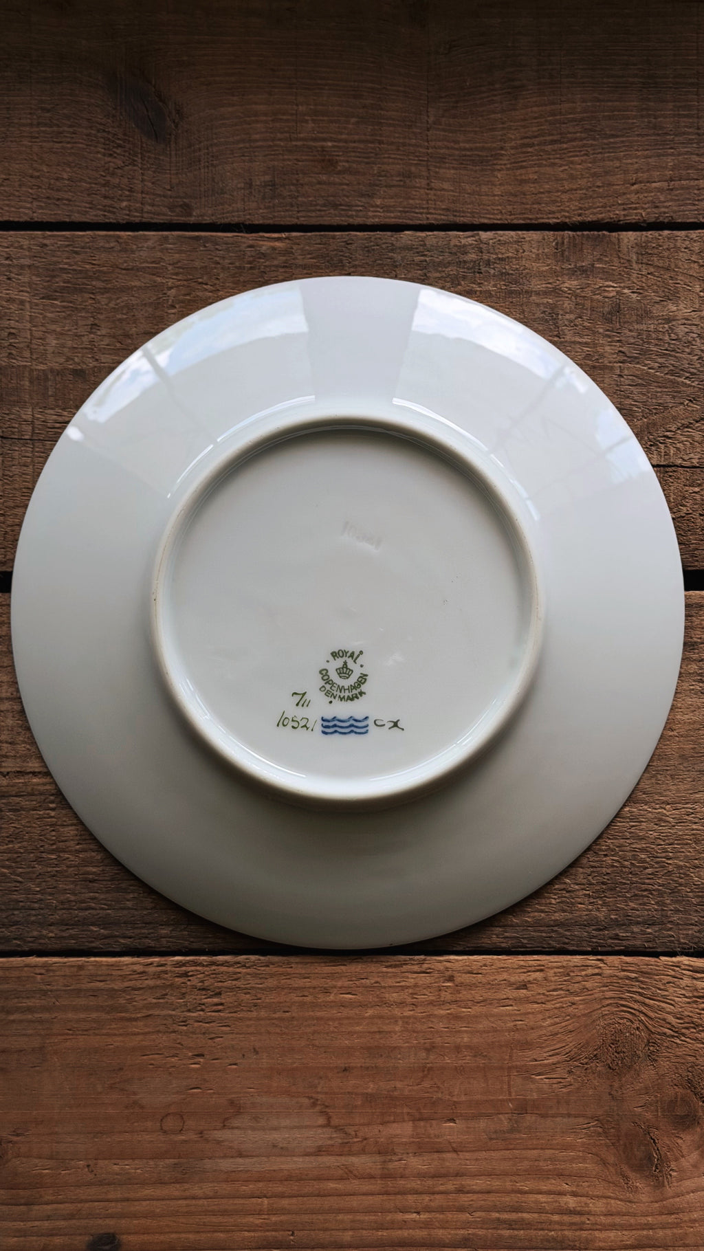 A Vintage Royal Copenhagen Brown Iris Salad Plate shown from the back, featuring a small blue and green printed mark near the center, placed on a dark brown wooden surface.