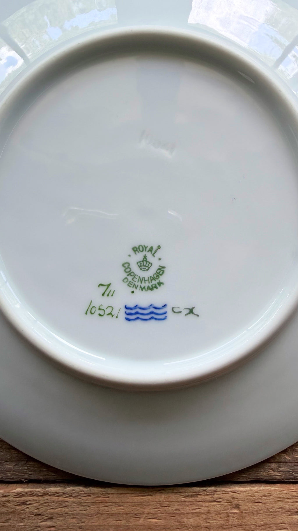 The underside of a white ceramic plate or dish, showing a green and blue Royal Crown Glass maker's mark with the text '7th 1052' on a wooden surface.
