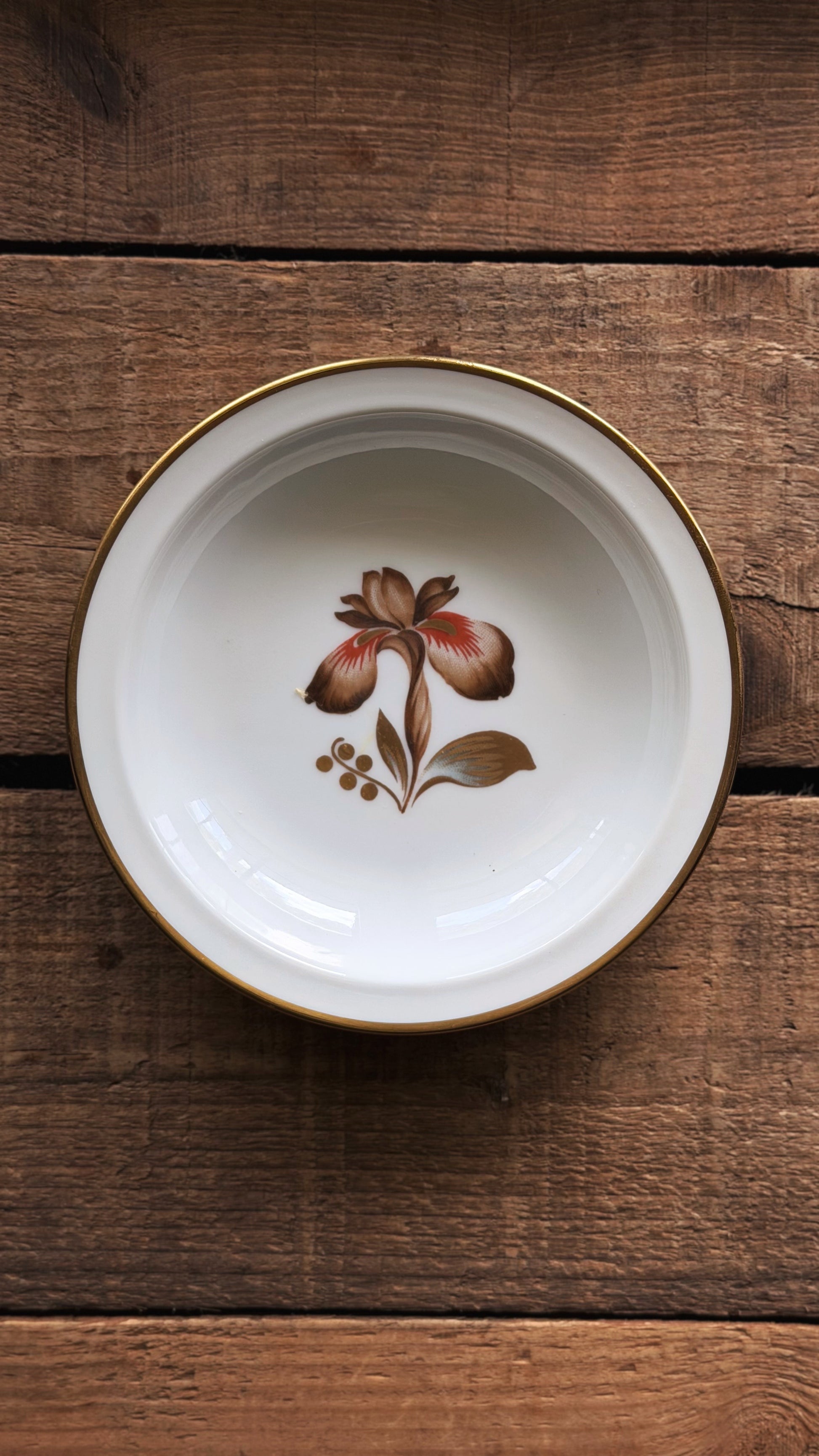 A Vintage Royal Copenhagen Brown Iris Fruit Bowl with a gold rim and a central floral illustration, photographed from above on a rustic dark wood background.