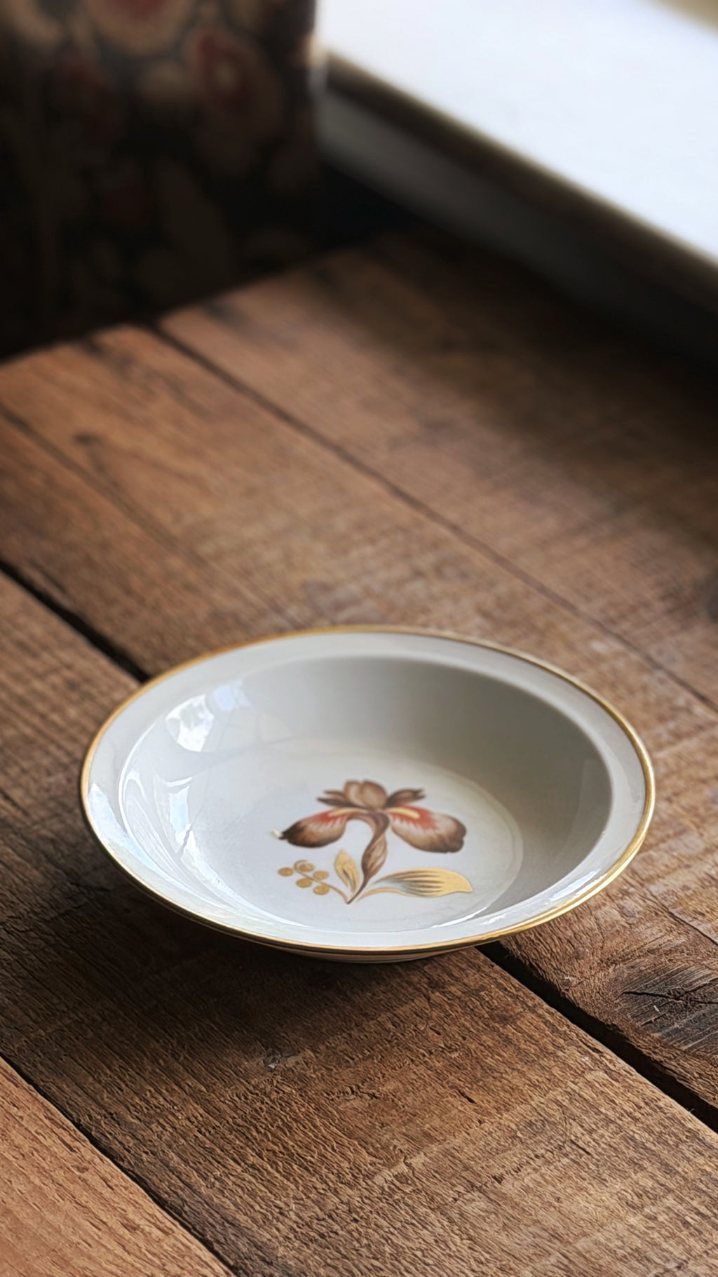A Vintage Royal Copenhagen Brown Iris Fruit Bowl with a gold rim and a delicate floral design in the center, resting on a rustic wooden table.
