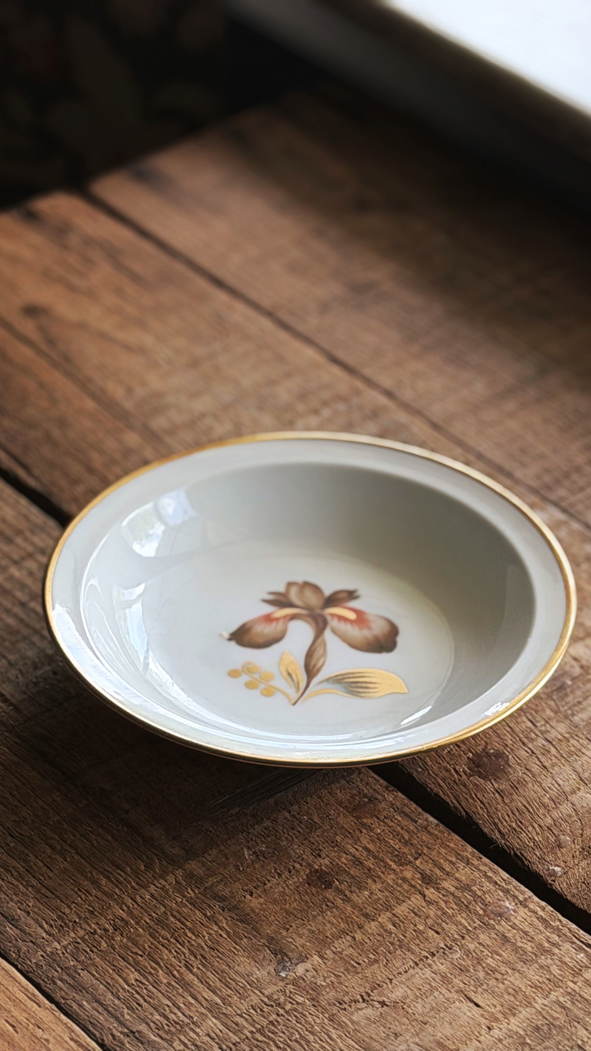 A Vintage Royal Copenhagen Brown Iris Fruit Bowl with a gold rim and a hand-painted iris flower in the center, resting on a rustic wooden surface.