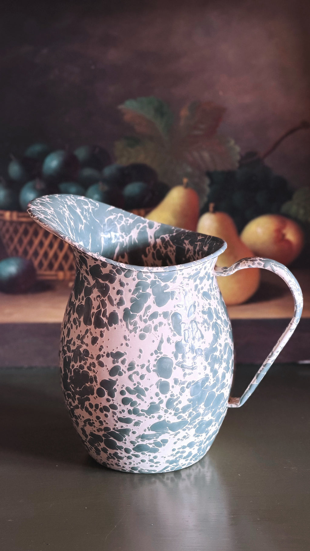 A vintage-style enamel pitcher with a speckled pattern of black, gray, and white on a pink base, set against a dark, rustic background with blurred fruit.