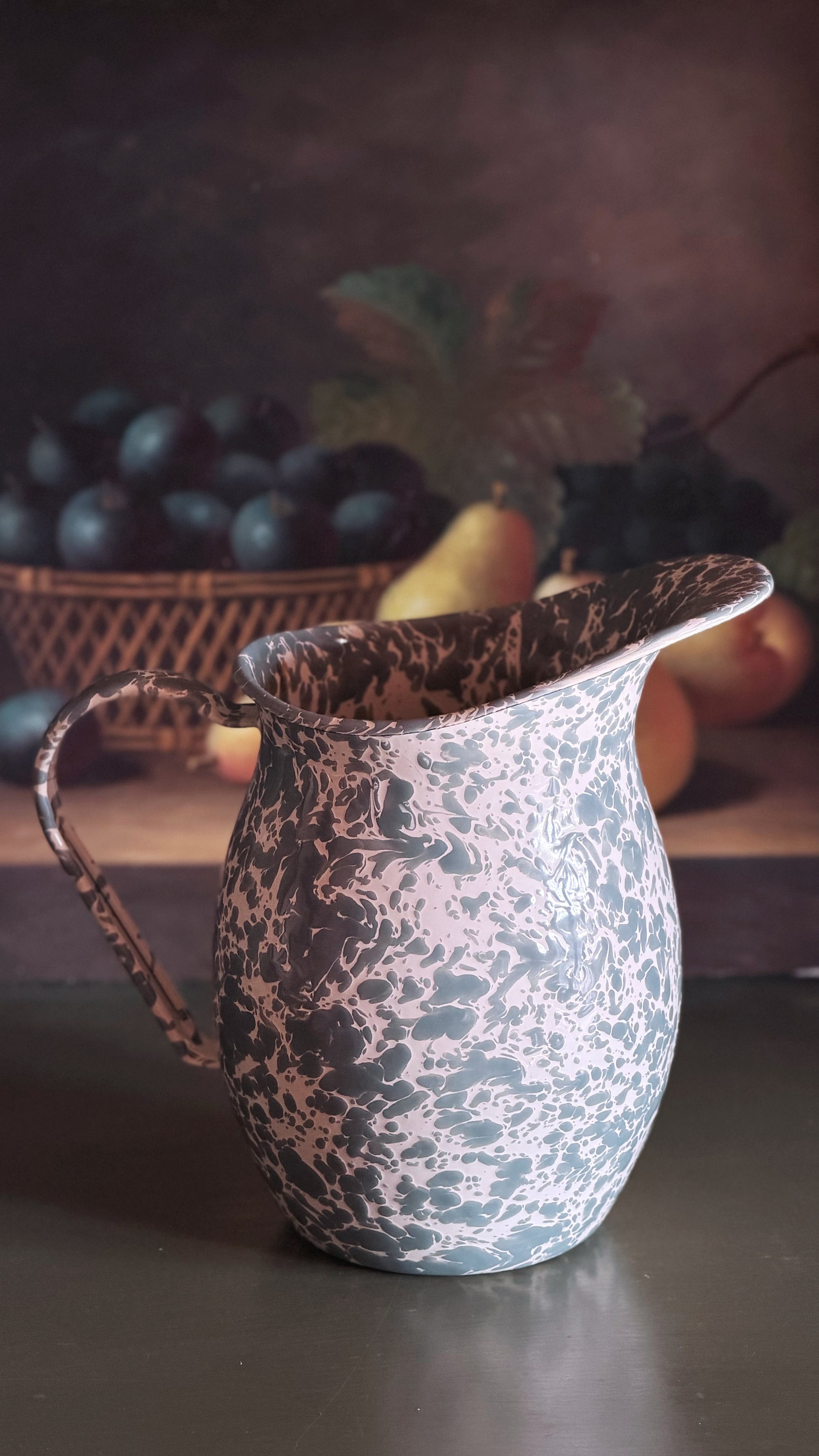 A vintage-style enamel pitcher with a mottled gray and white marbled pattern, set on a dark tabletop with a blurred rustic fruit basket in the background.