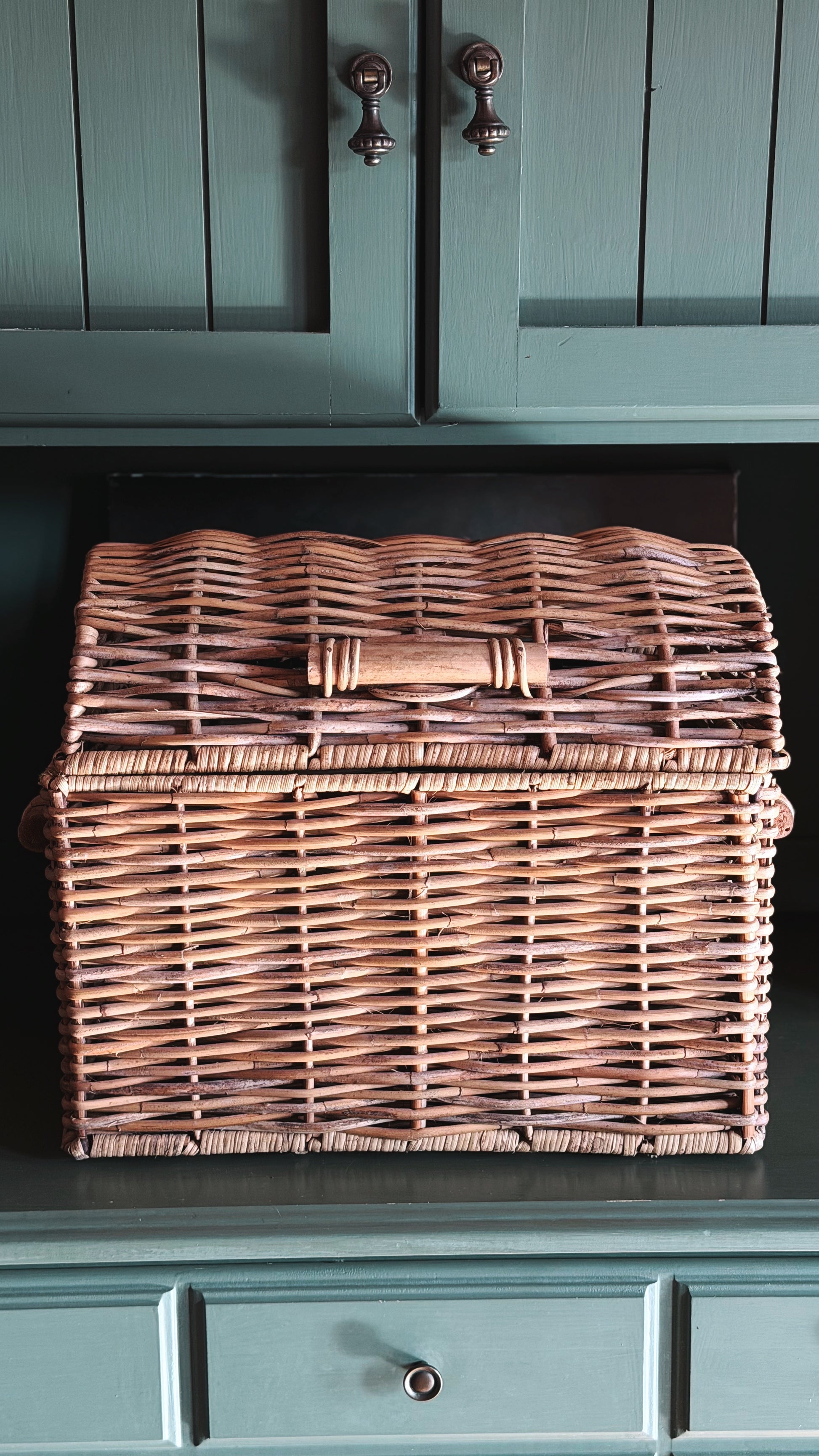 A rectangular woven wicker storage basket with a lid and central handle, displayed on a dark teal cabinet shelf.