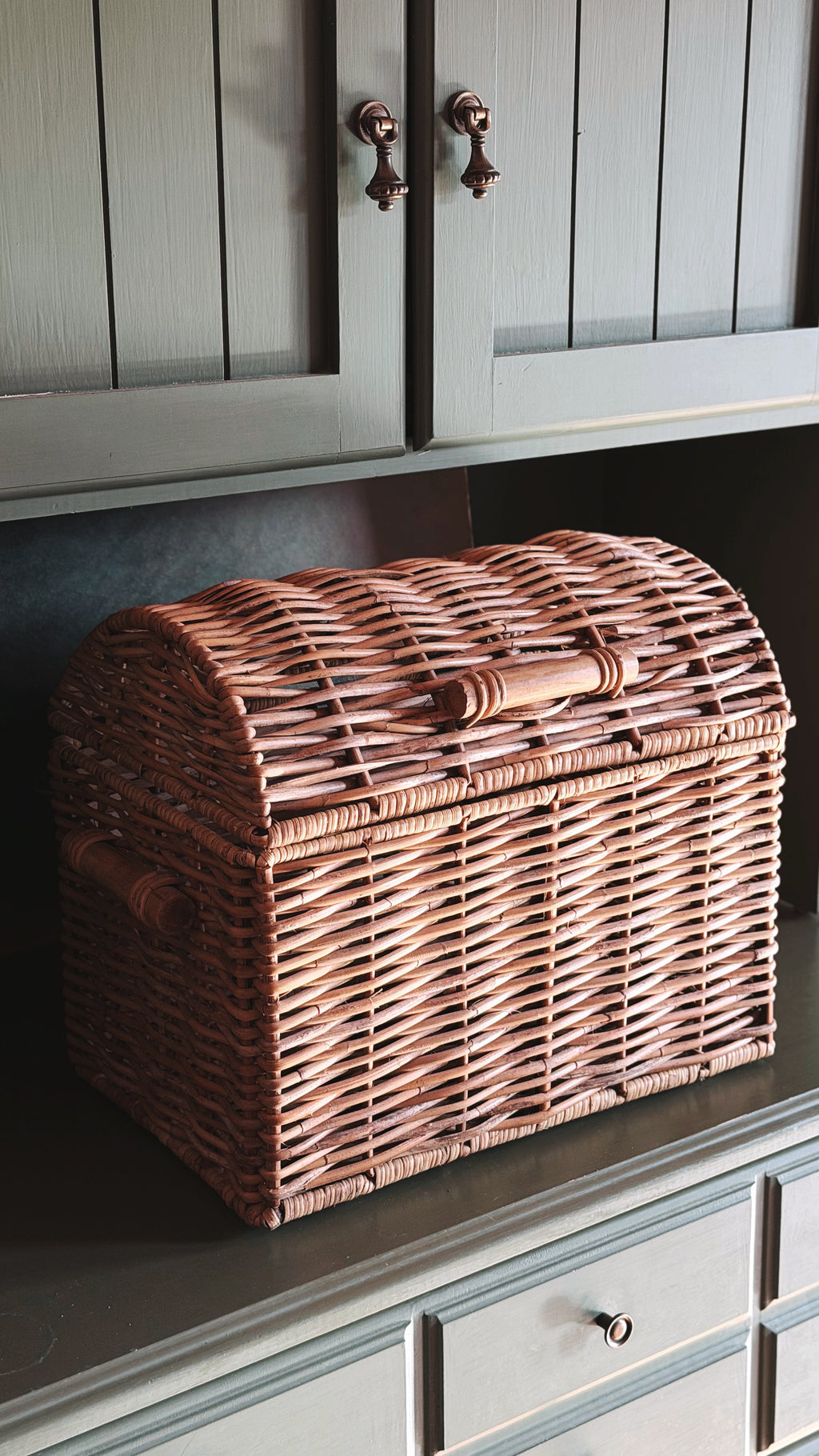 A rustic wicker chest with a hinged lid and wooden handle, displayed on a dark countertop beneath gray cabinets.