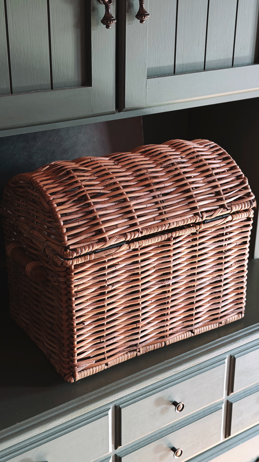 A brown woven wicker chest with a hinged lid sits on a dark gray cabinet shelf in a home setting.