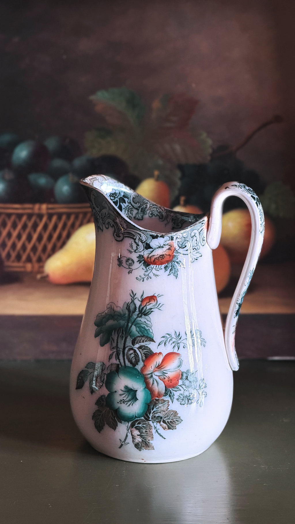 An Antique Francis Morley Polychrome Ironstone Pitcher decorated with a colorful floral pattern featuring green leaves, red berries, and blue-green grapes, set against a dark, rustic background with blurred fruit.