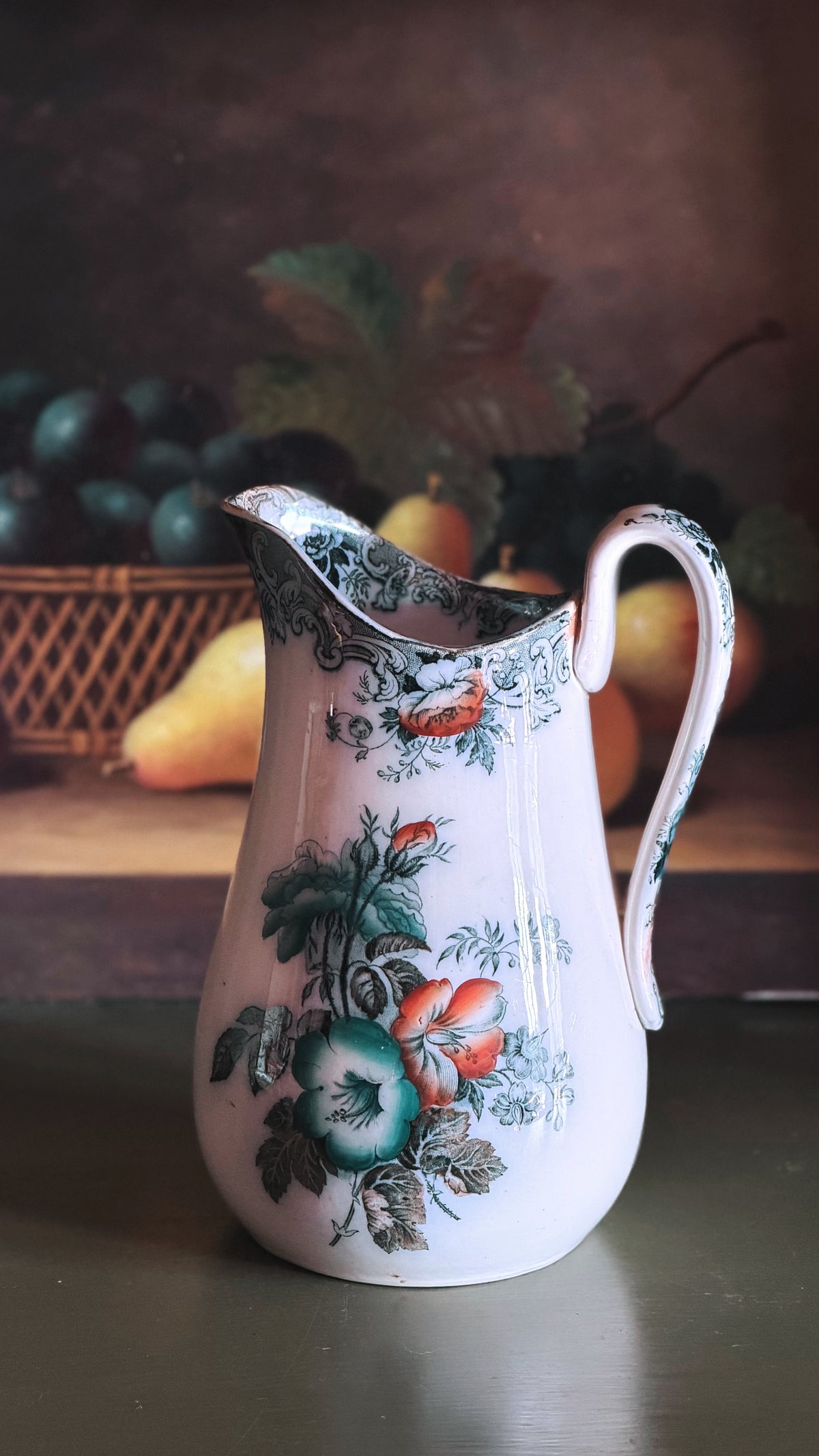 An Antique Francis Morley Polychrome Ironstone Pitcher decorated with a colorful floral pattern featuring green leaves, red berries, and blue-green grapes, set against a dark, rustic background with blurred fruit.