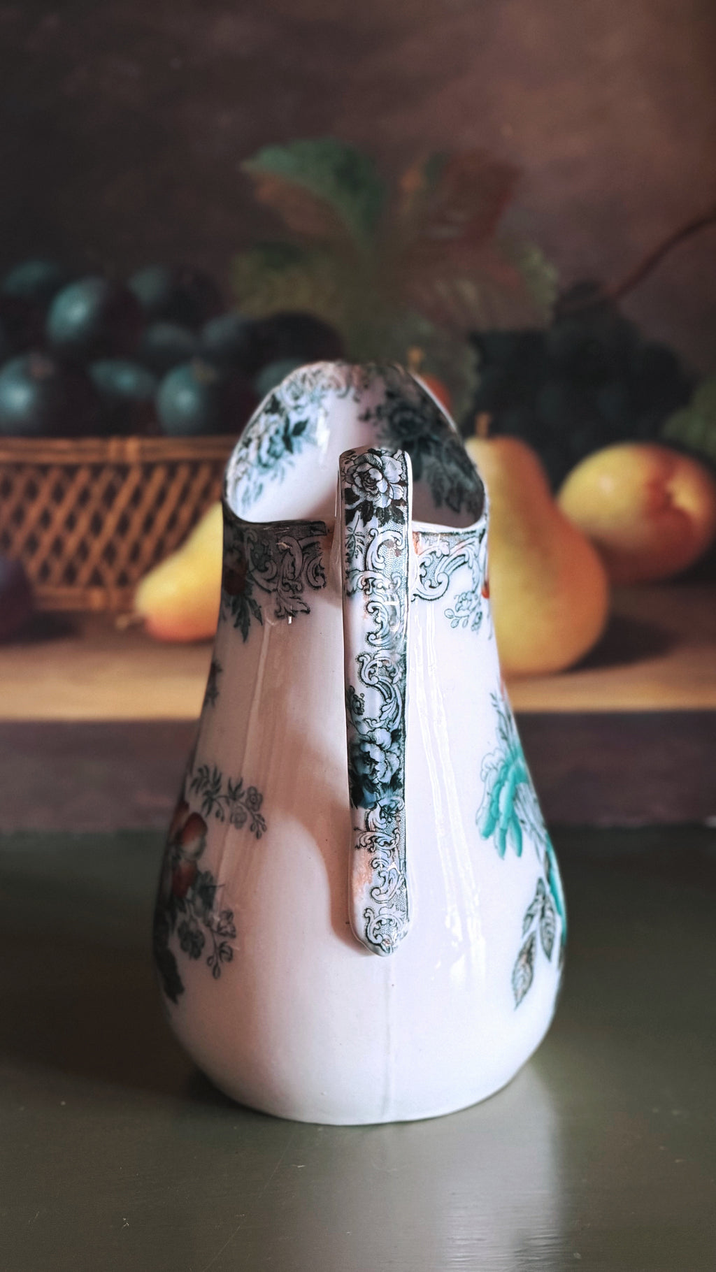 An Antique Francis Morley Polychrome Ironstone Pitcher with green and blue floral patterns and a silver handle, set against a dark rustic background with blurred fruit.
