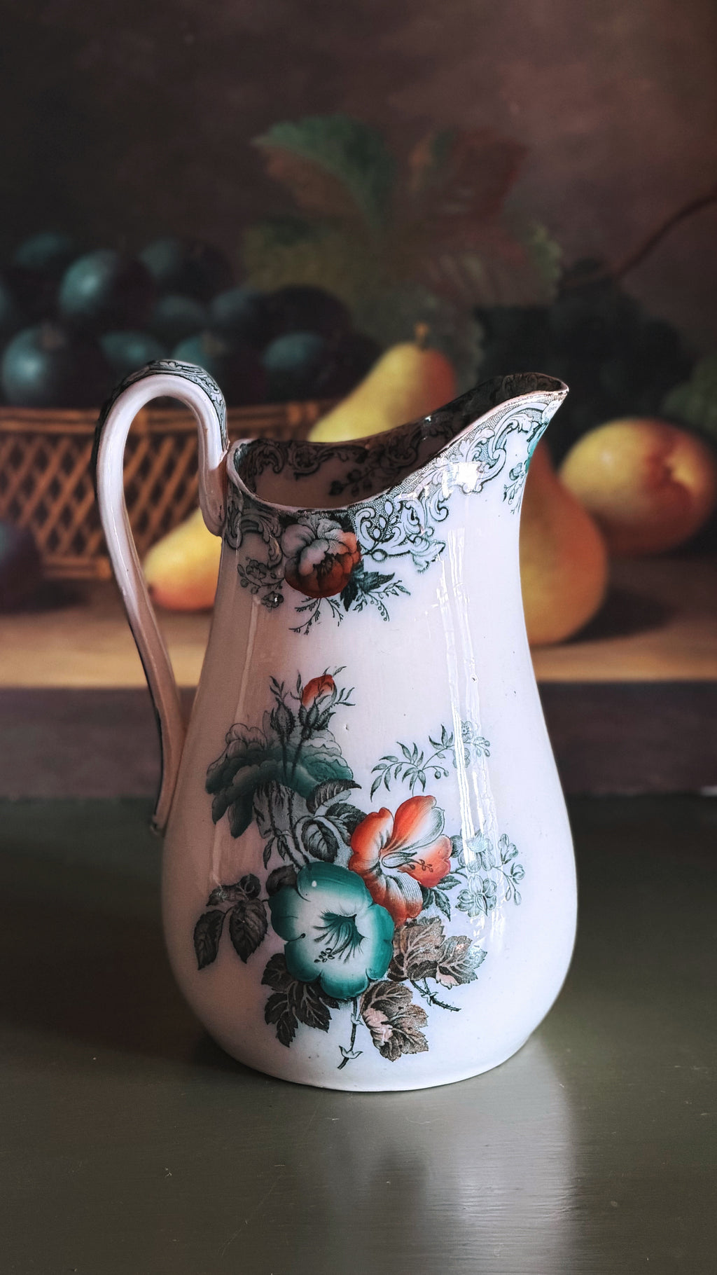 An Antique Francis Morley Polychrome Ironstone Pitcher decorated with hand-painted floral motifs in green, orange, and blue, set against a dark, rustic background with blurred fruit.