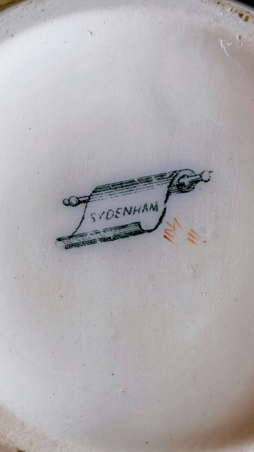 Close-up of the inside bottom of a white ceramic plate or dish, showing a printed brand mark reading SYDENHAM with a small orange monogram.