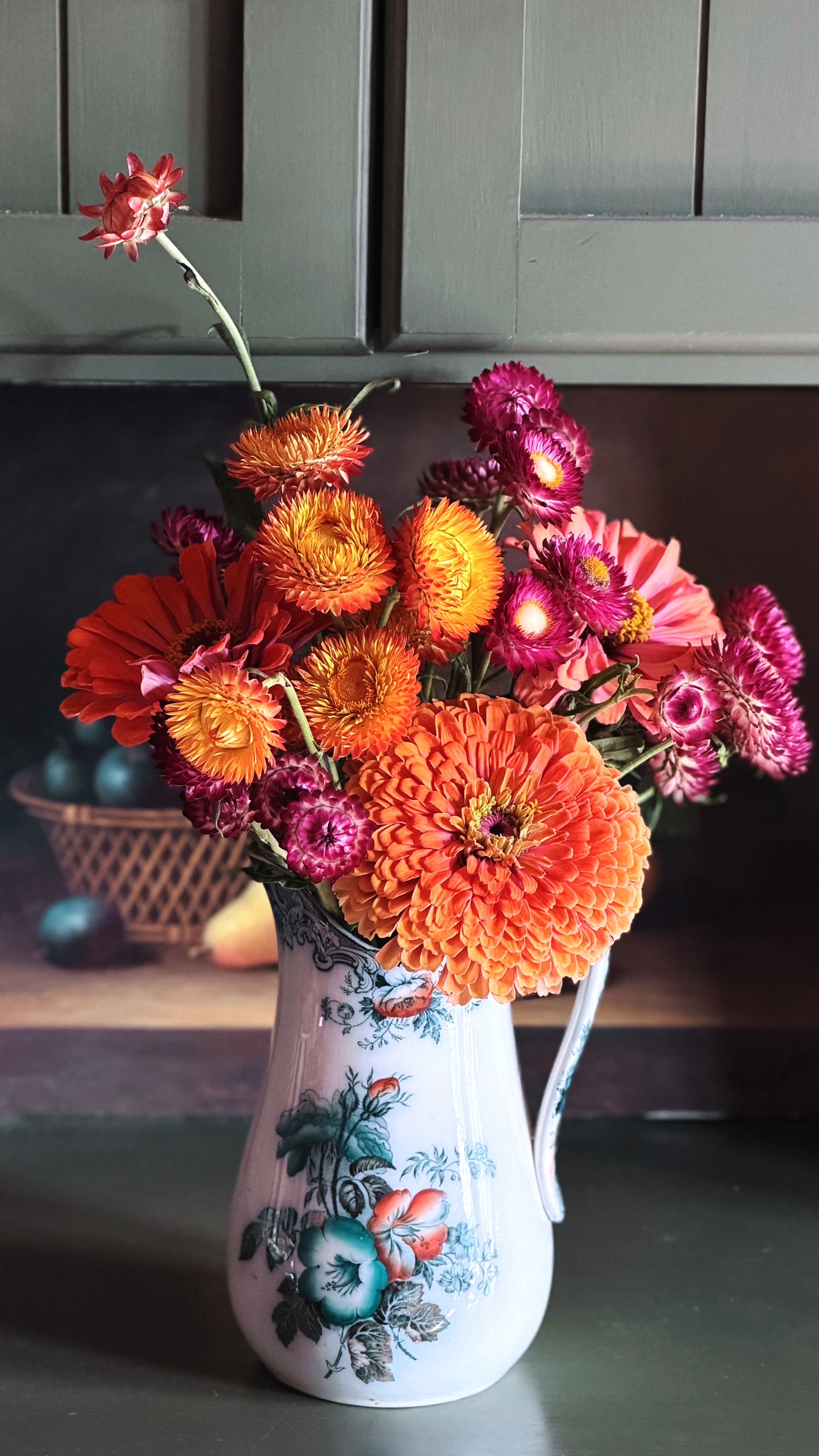 A vibrant floral arrangement of orange, pink, and red flowers in a white ceramic pitcher vase with blue and green floral patterns, set on a dark countertop with a dark wood cabinet background.
