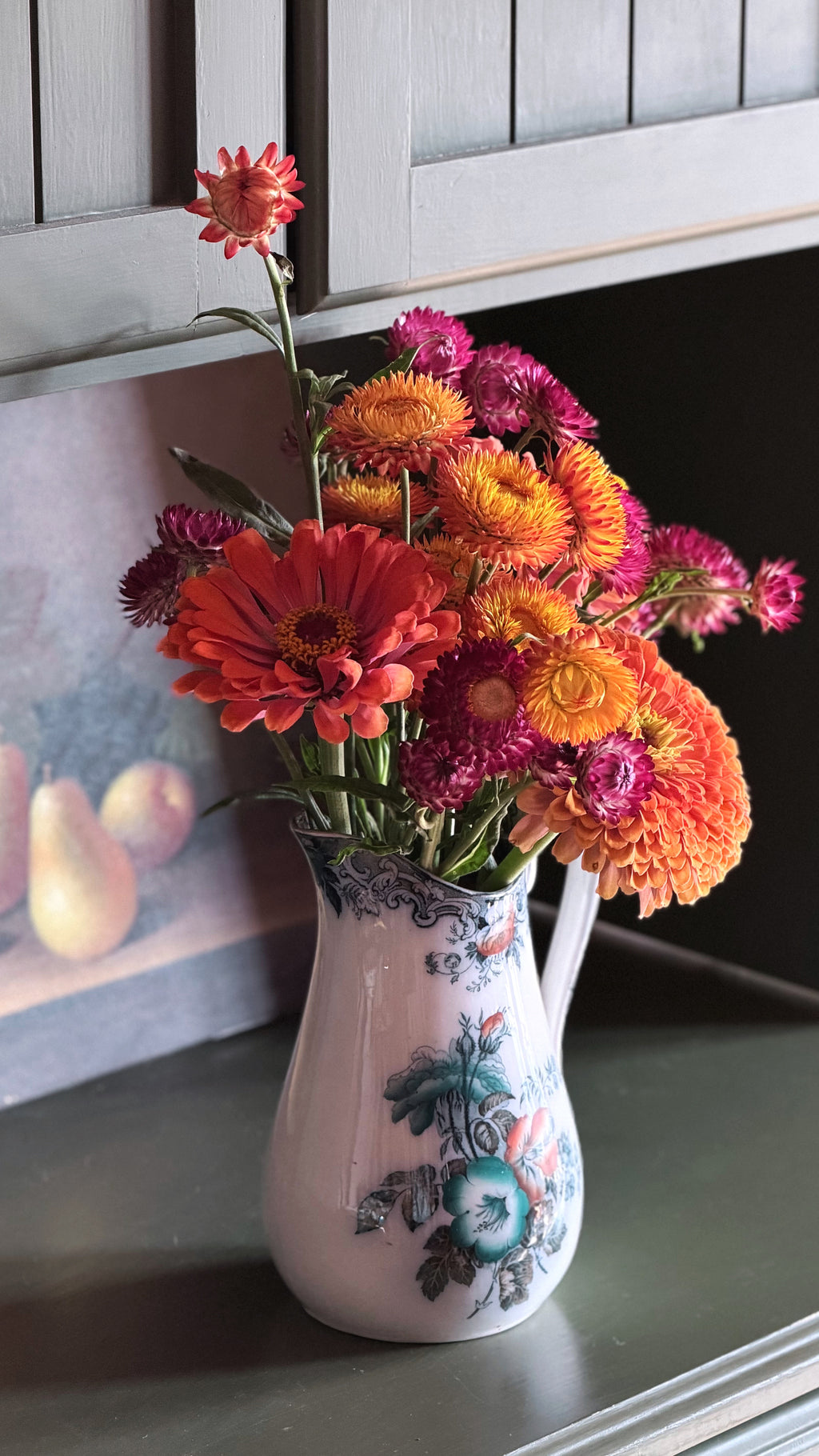 An ironstone pitcher vase with a white base and blue floral illustration, filled with a colorful bouquet of orange, pink, and yellow strawflowers, set on a dark countertop.