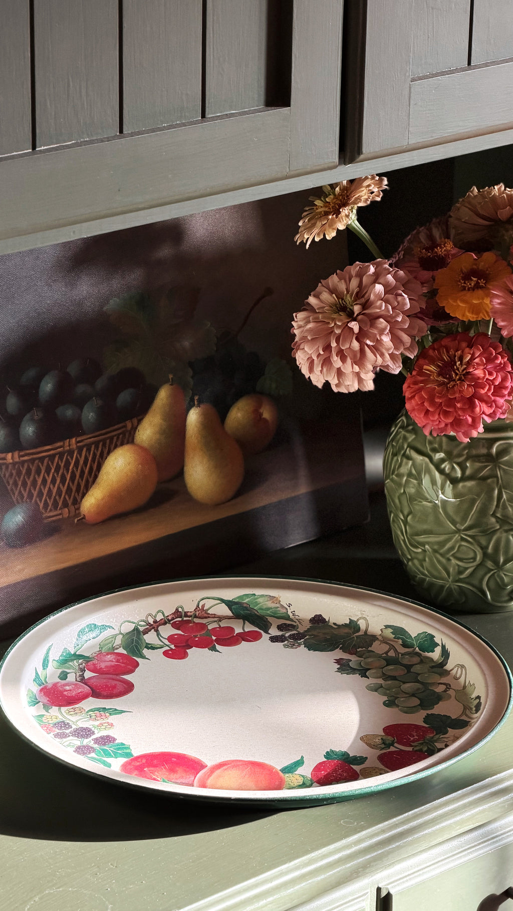 A Vintage Potpourri Press Floral Round Metal Serving Tray decorated with a red cherry and green grapevine border, displayed on a dark countertop next to a floral arrangement and a fruit-themed wall mural.