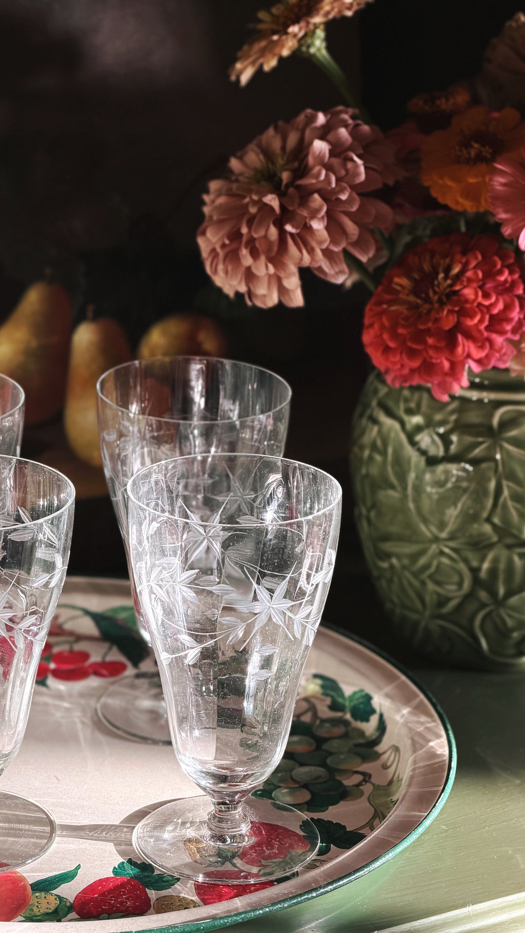 Clear etched glass iced tea glasses on a decorative tray with a floral rim, set in a softly lit interior scene with flowers and a green textured vase in the background.