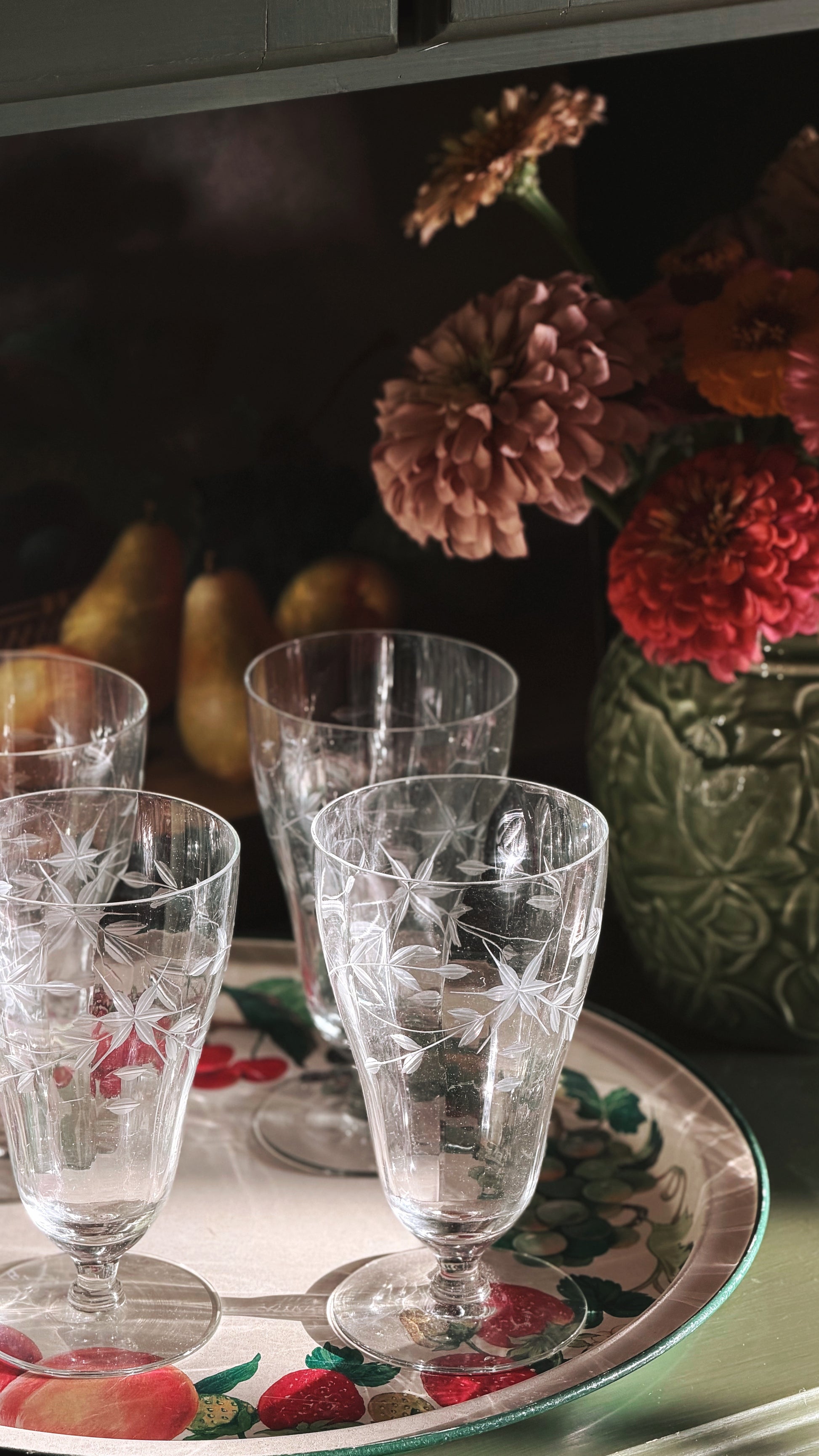 Four clear cut-glass wine glasses with etched starburst patterns arranged on a decorative tray with fruit illustrations, set in a dimly lit interior with flowers and pears in the background.