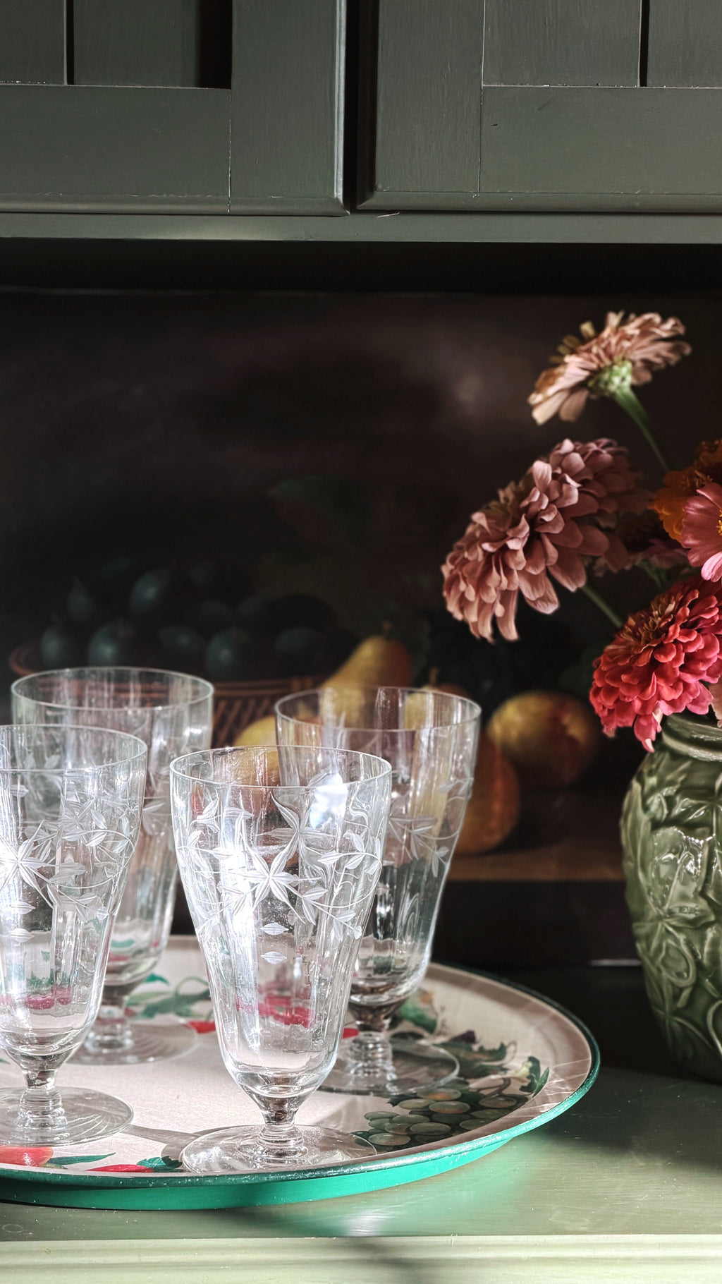 A set of clear cut-crystal glasses with etched star patterns, arranged on a decorative round tray with a green rim, set against a dark rustic background with flowers and pears.