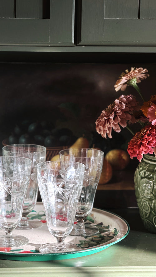 A set of clear cut-crystal glasses with etched star patterns, arranged on a decorative round tray with a green rim, set against a dark rustic background with flowers and pears.