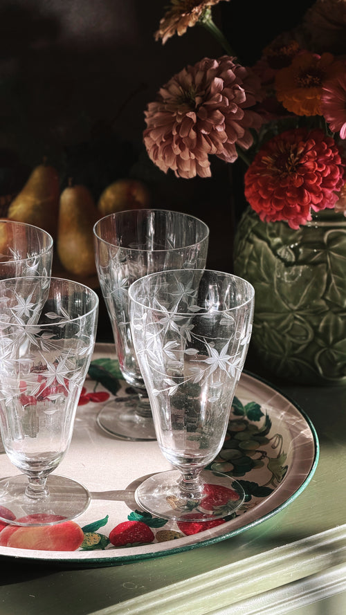 Four clear cut-crystal wine glasses with etched floral star patterns, arranged on a decorative tray with strawberries and fruit, with flowers in the background.