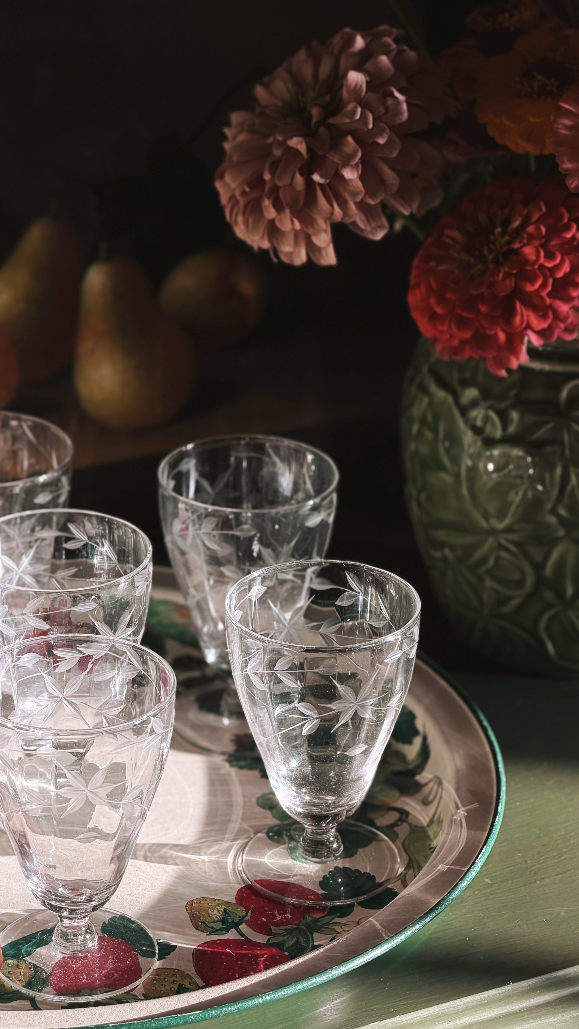 A set of Vintage Etched Crystal Starburst Aperitif | Cordial Glasses arranged on a decorative floral tray, with a dark green textured vase of flowers in the background.
