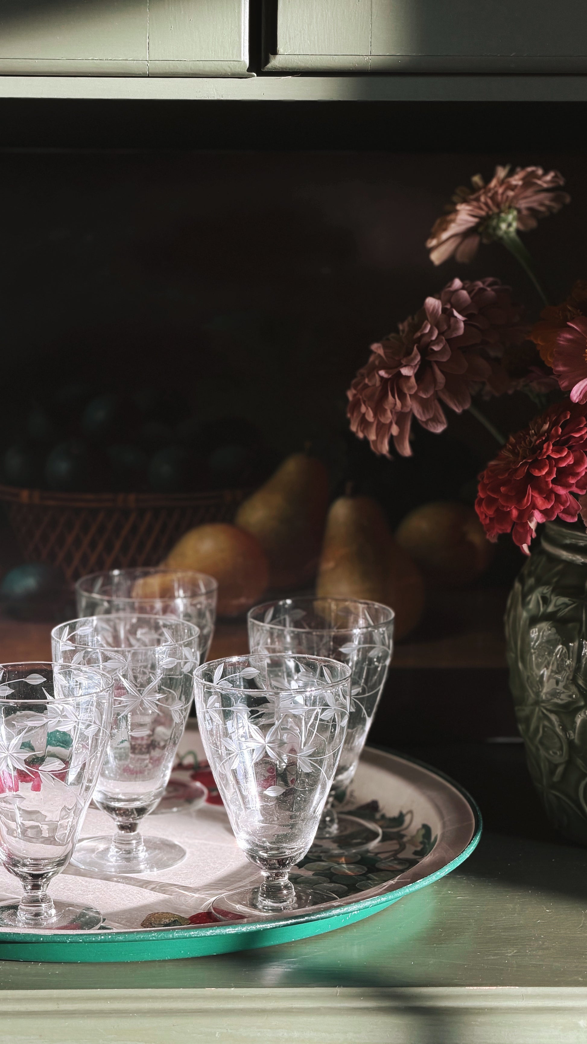 A set of Vintage Aperitif | Cordial Glasses with etched floral patterns arranged on a decorative tray, set against a dark, moody background with pears and flowers.