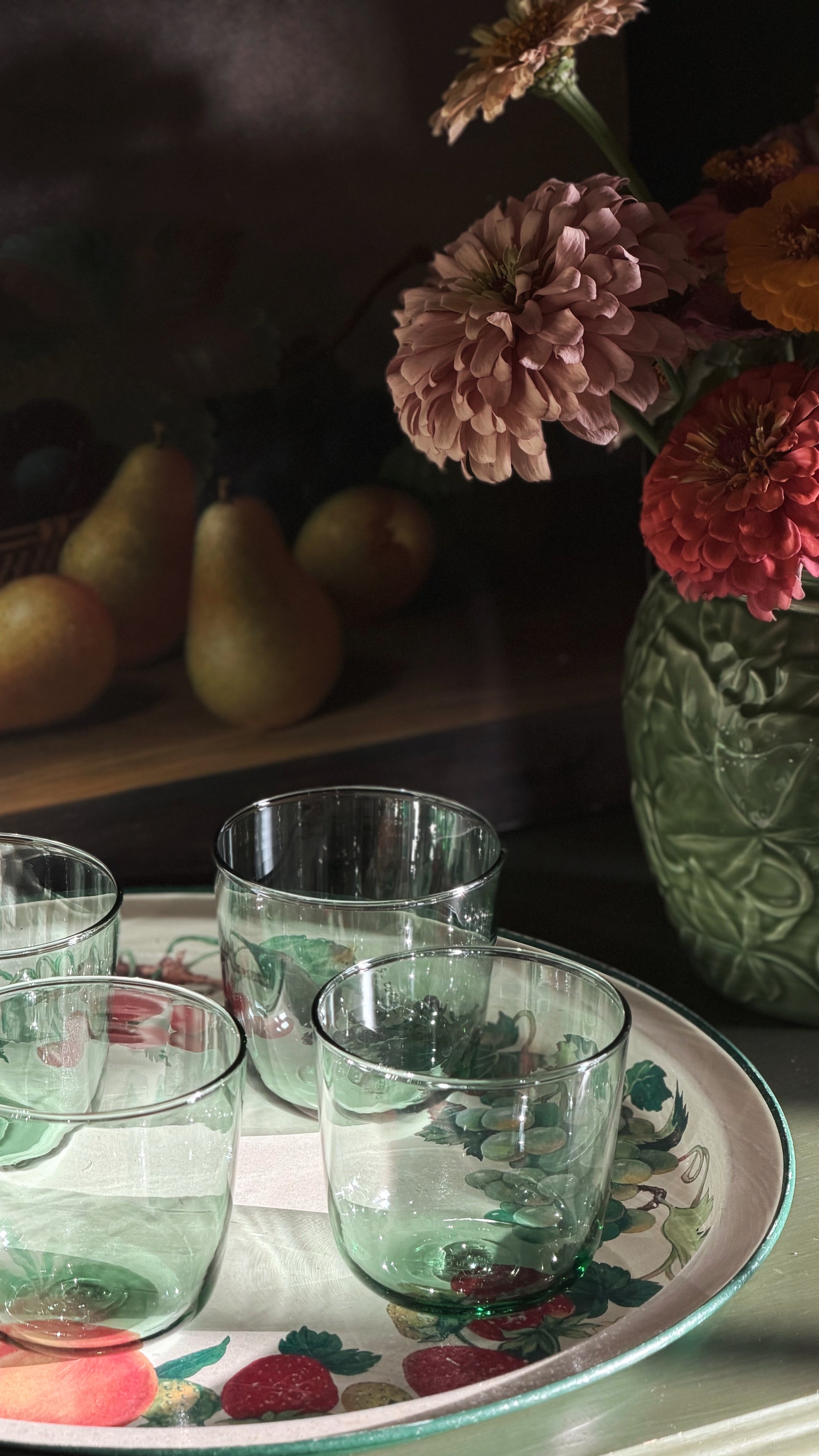 A set of glass tumblers with a subtle green tint, arranged on a decorative floral tray next to a green embossed ceramic vase filled with flowers, with pears blurred in the background.