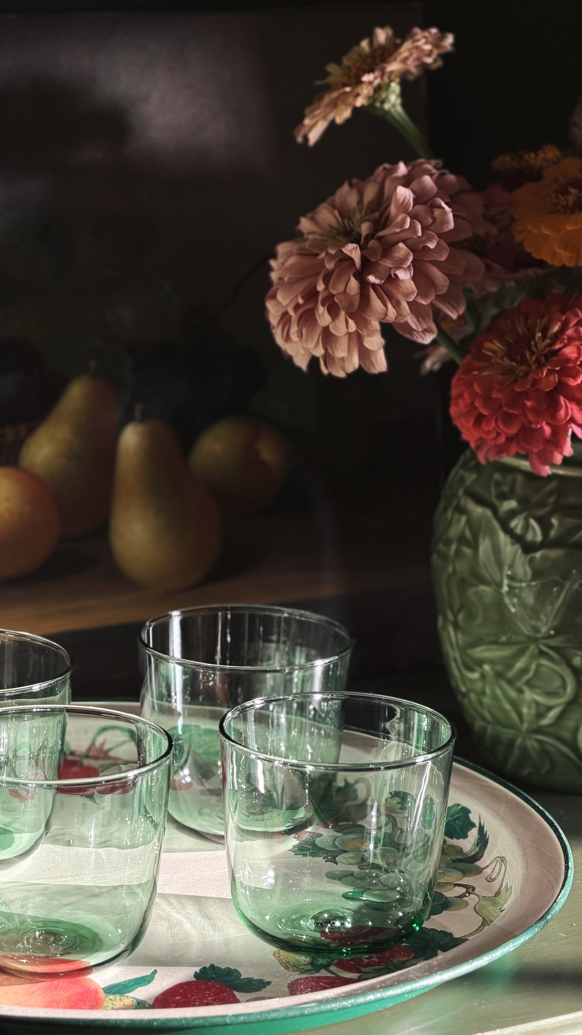 Vintage IKEA France Green Glass Tumblers arranged on a decorative floral tray, with a dark green embossed vase of flowers and pears in the softly lit background.