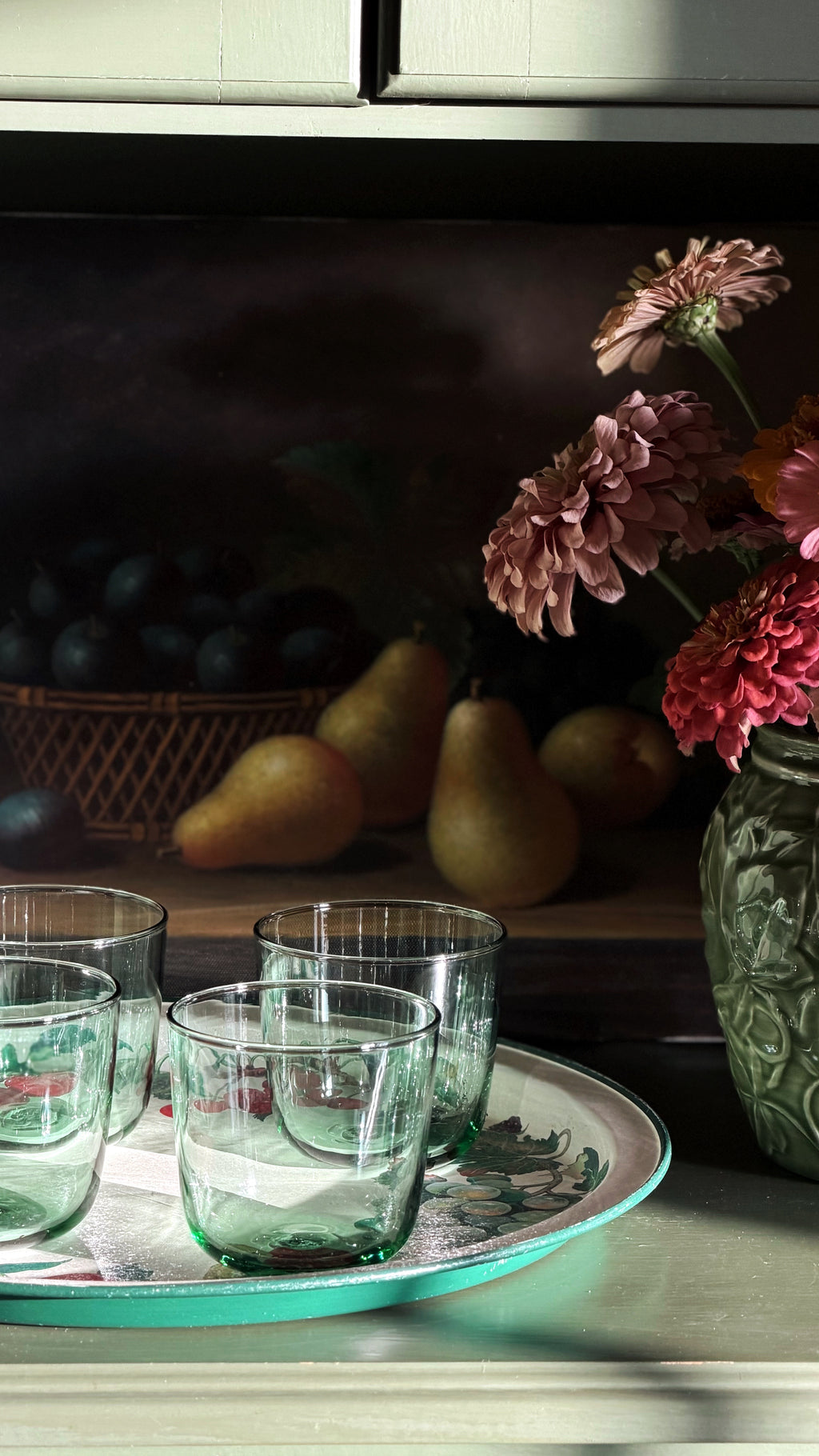 A still life scene with Vintage IKEA France Green Glass Tumblers on a round tray, set against a dark painted background with pears, a basket of dark fruit, and pink flowers in a textured vase.