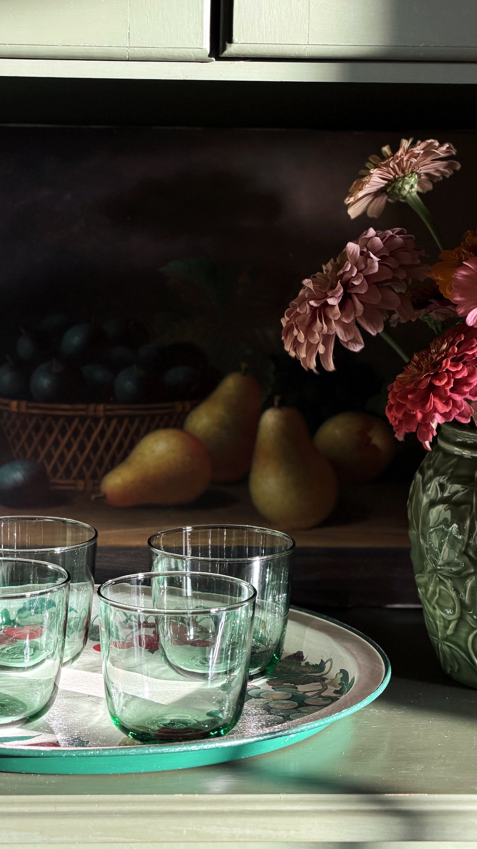 A still life scene with Vintage IKEA France Green Glass Tumblers on a round tray, set against a dark painted background with pears, a basket of dark fruit, and pink flowers in a textured vase.