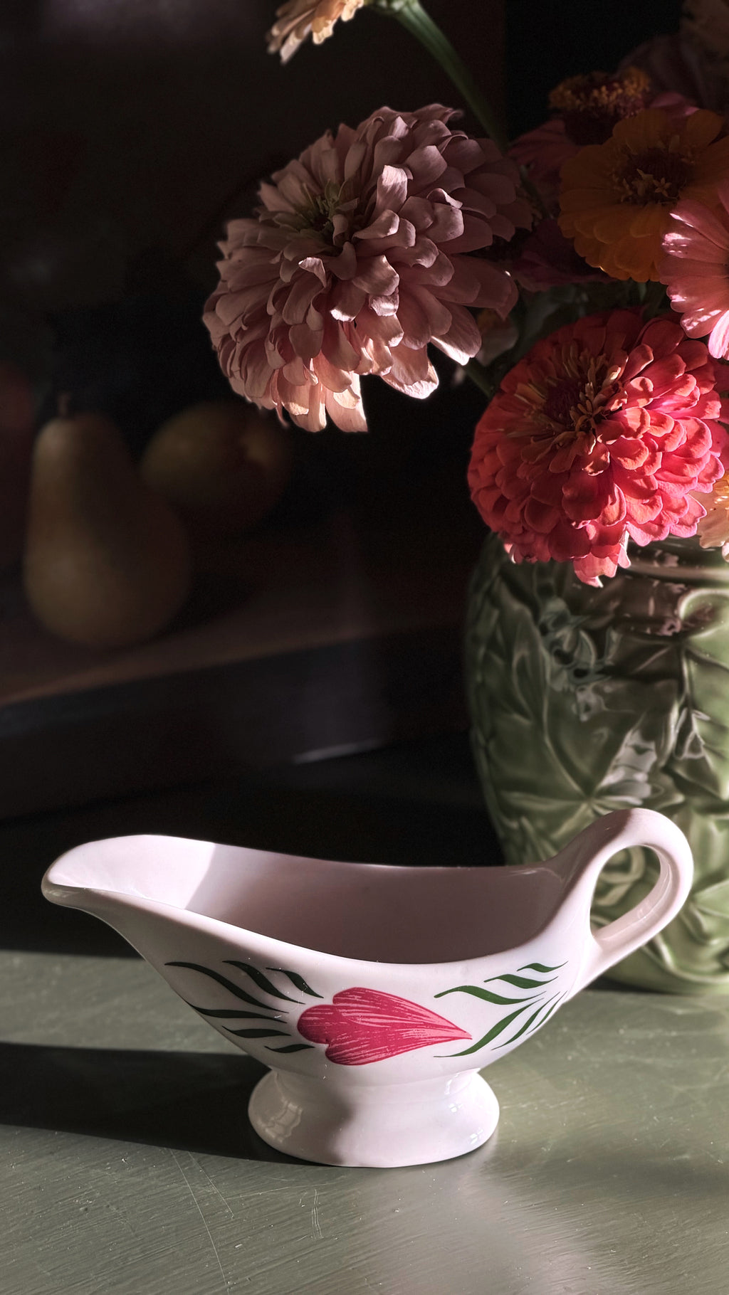 A white ceramic gravy boat with a pink floral design sits on a tabletop in warm, dramatic sunlight, with a vase of flowers blurred in the background.