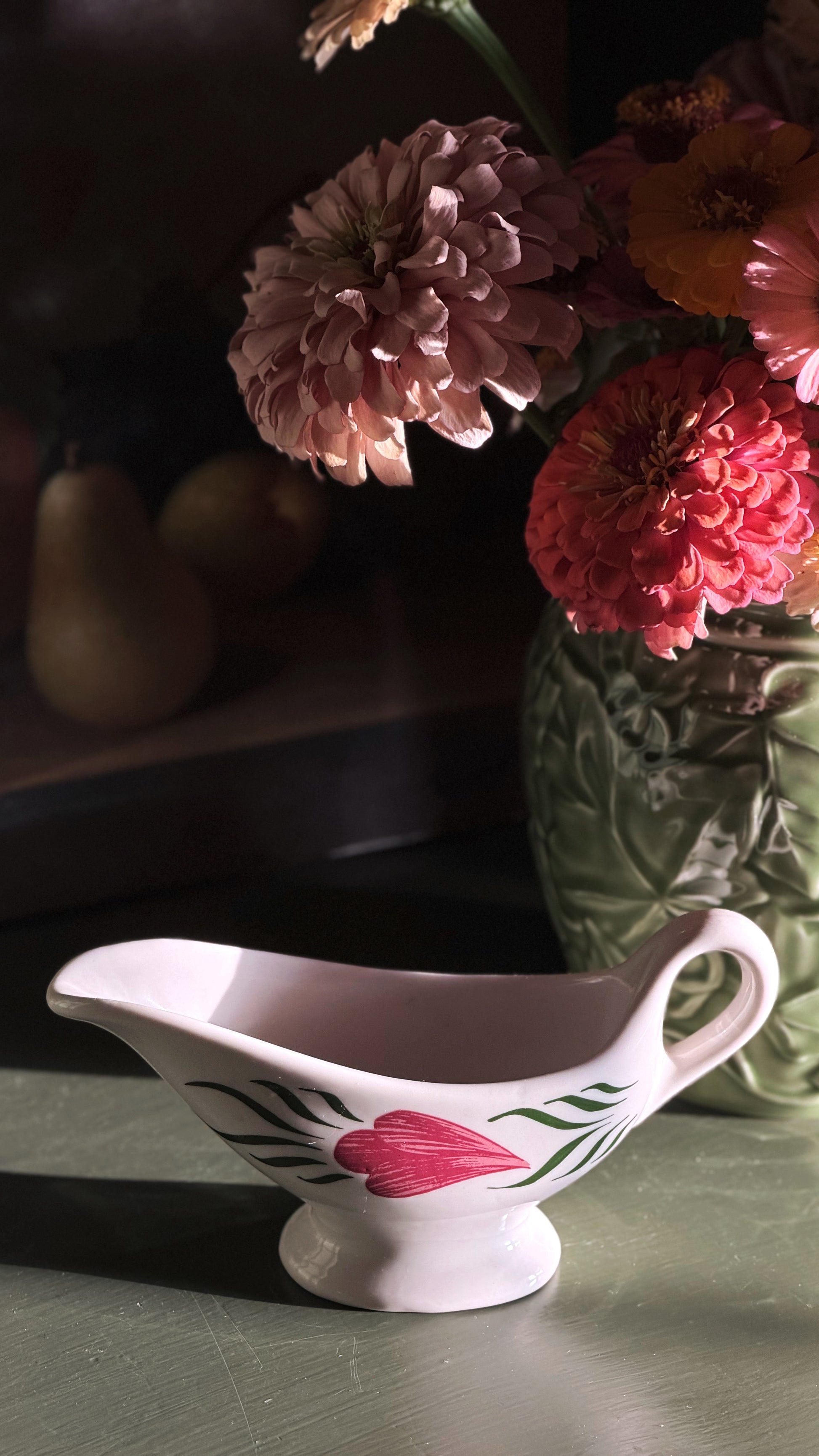A white ceramic gravy boat with a pink floral design sits on a tabletop in warm, dramatic sunlight, with a vase of flowers blurred in the background.