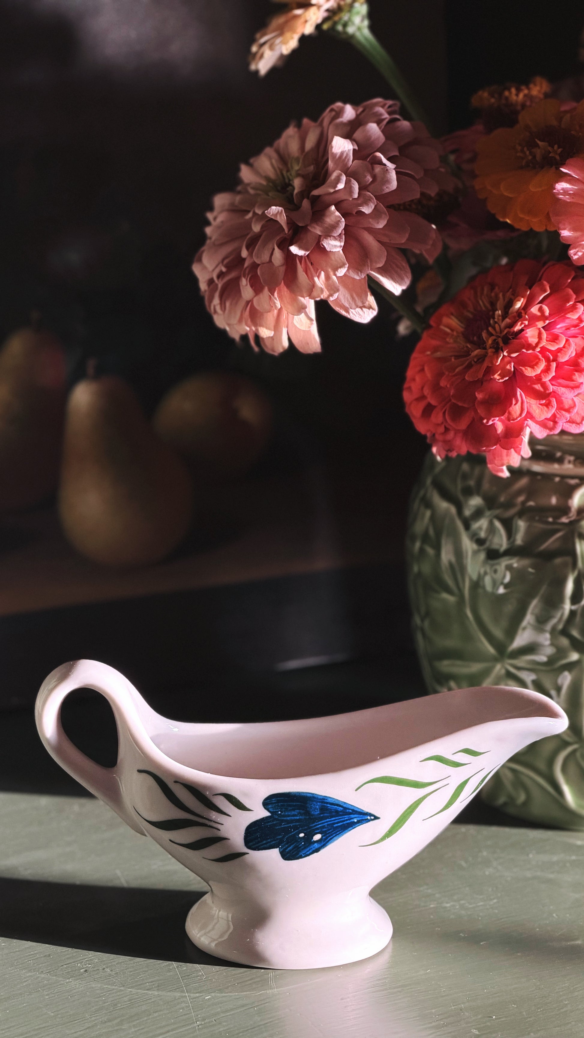 A lVintage Syracuse China Gravy Boat with a blue and green floral design, set on a tabletop next to a vase of pink and red flowers in a dimly lit interior.