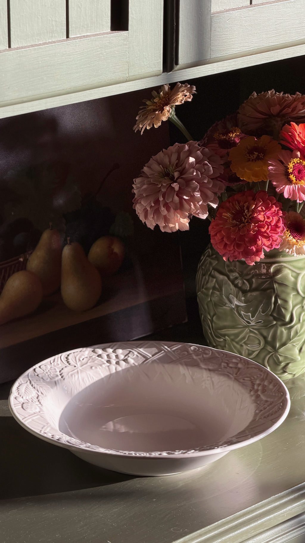 A Mikasa English Countryside bowl with embossed floral detailing around the rim, set on a tabletop next to a green textured vase filled with pink, red, and orange flowers.