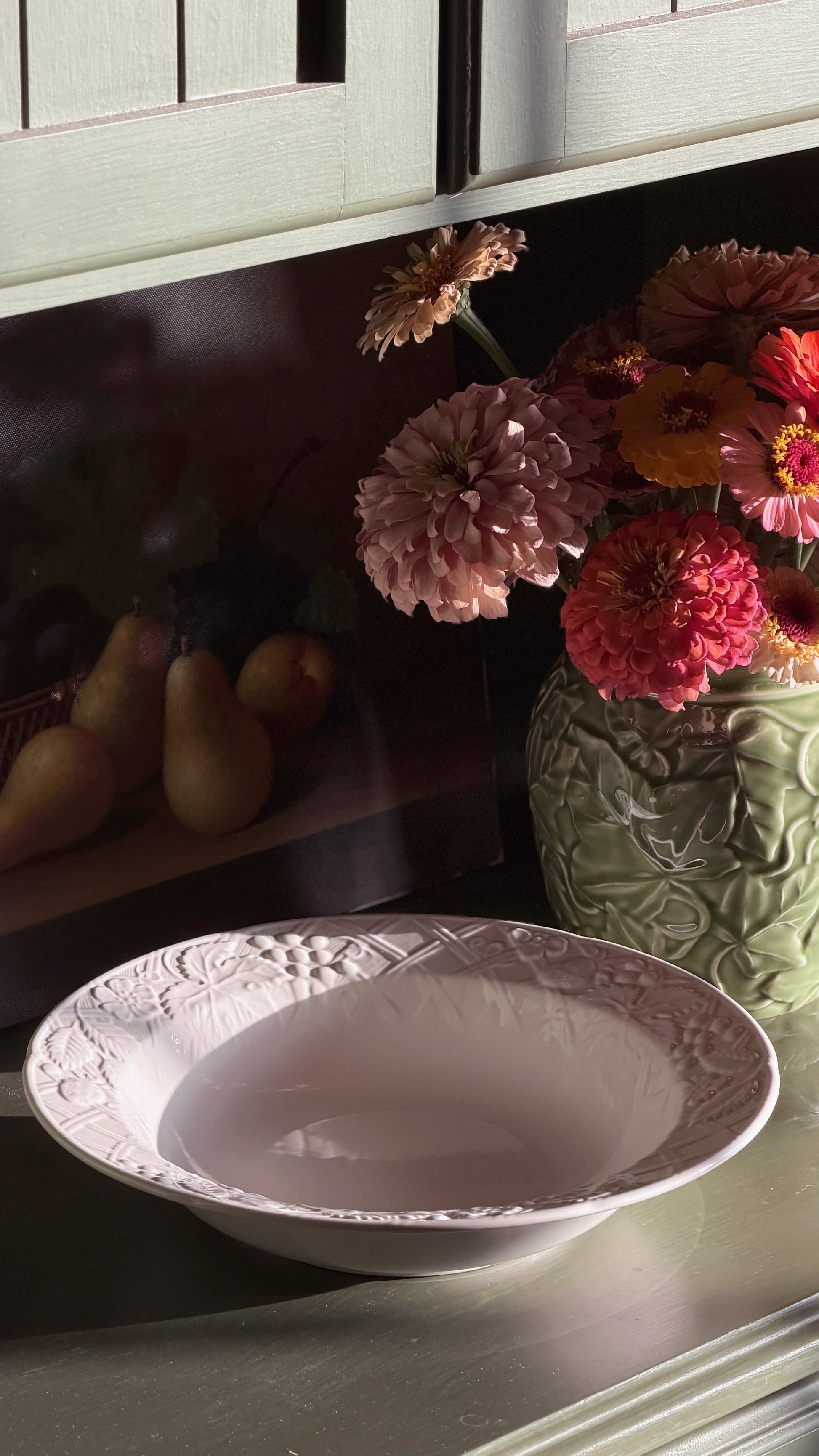 A Mikasa English Countryside bowl with embossed floral detailing around the rim, set on a tabletop next to a green textured vase filled with pink, red, and orange flowers.