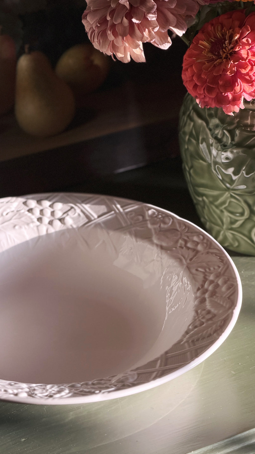 A close-up of a Mikasa English Countryside bowl with embossed floral patterns around the rim, set on a reflective surface with a green vase of flowers in the background.