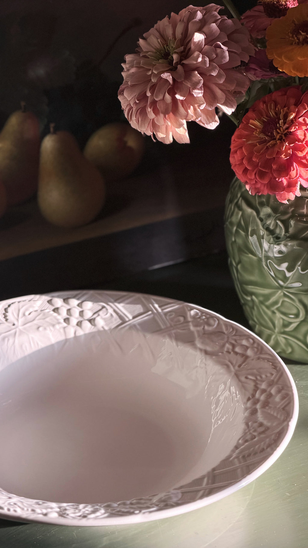 A white Mikasa English Countryside bowl with an embossed floral border, shown in a styled tabletop setting with pink flowers in a green vase and pears in the background.