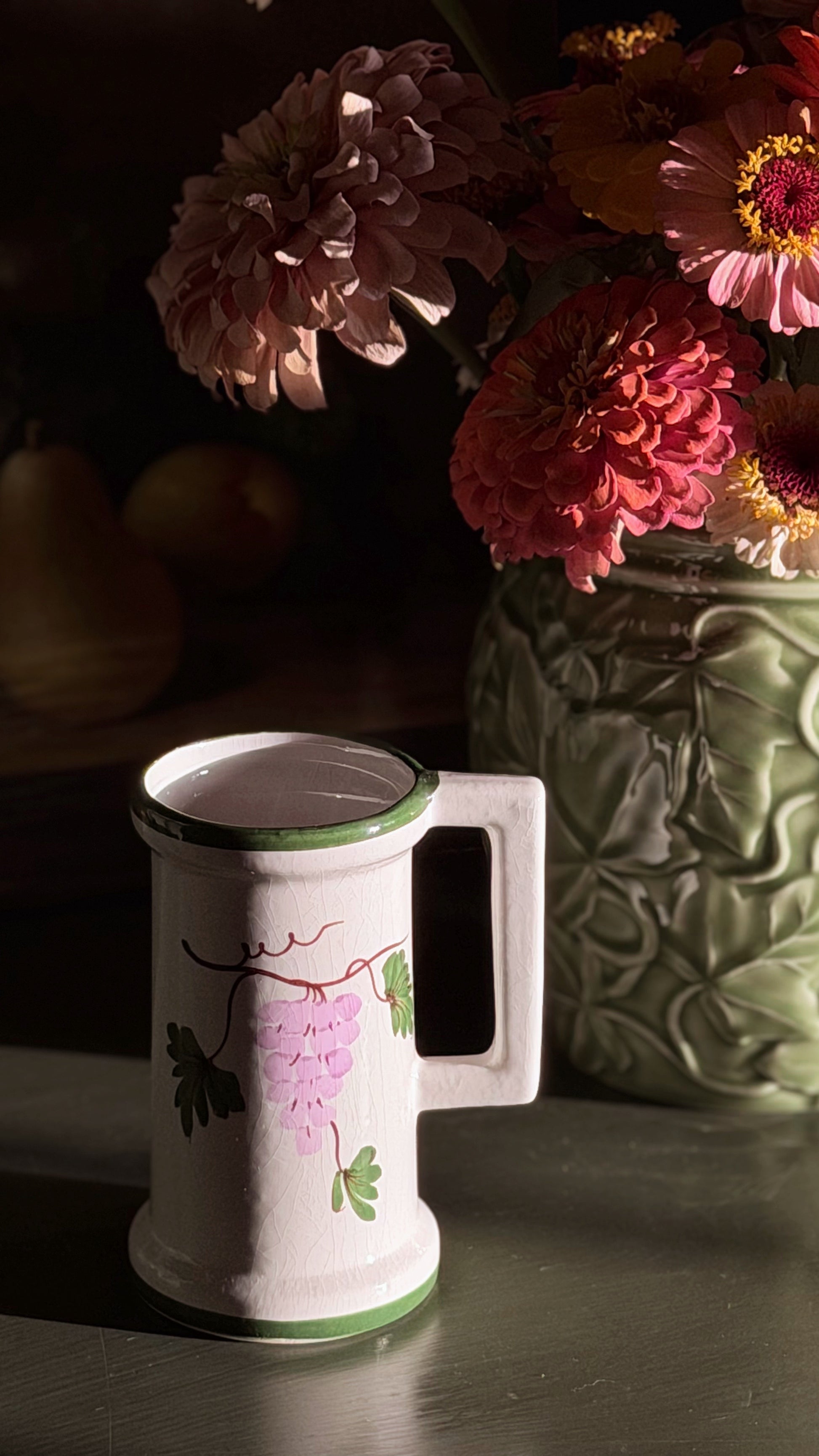 A Vintage Hand Painted Floral Ceramic Vessel with a green rim and handle, decorated with a pink grapevine illustration, set on a dark table next to a vase of pink and red flowers.