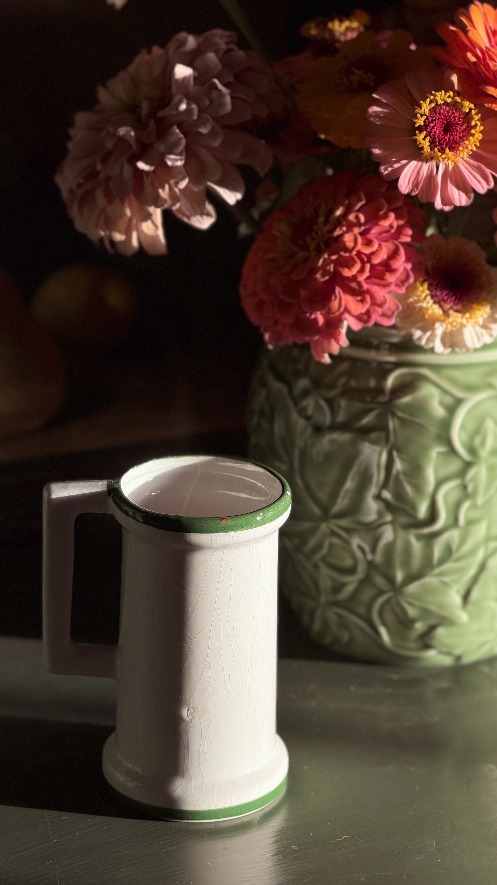 A Vintage Hand Painted Floral Ceramic Vessel with a green rim and base detail, set on a dark tabletop next to a green textured ceramic vase filled with pink and orange flowers.