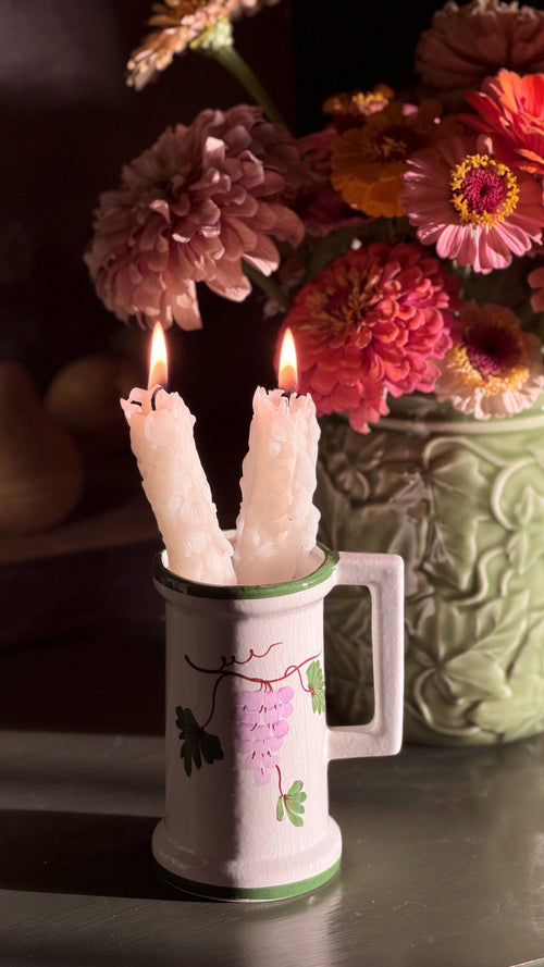 A Vintage Hand Painted Floral Ceramic Vessel with a green rim and floral decoration, holding two lit white taper candles, set against a dark background with a vase of colorful flowers.