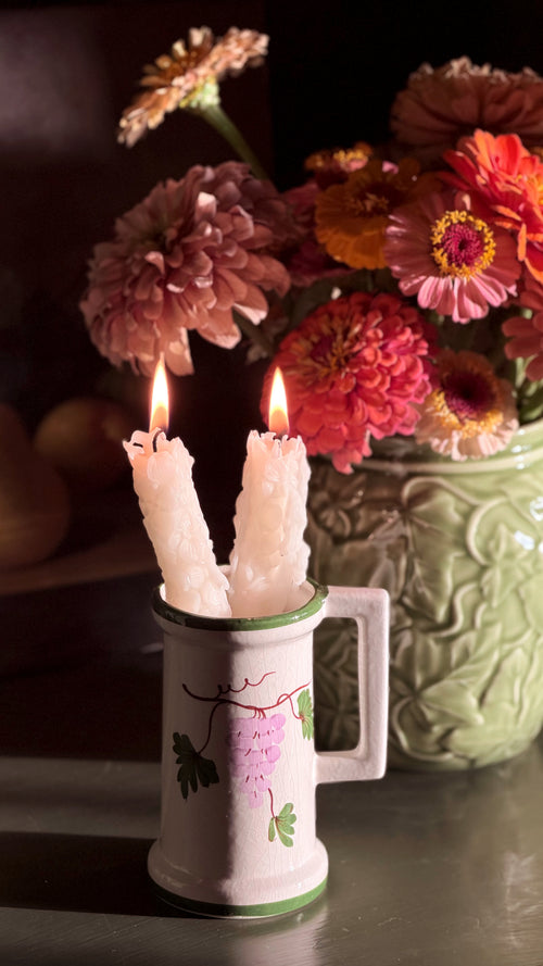 A Vintage Hand Painted Floral Ceramic Vessel with a green rim and floral print, holding two lit white taper candles. The mug is set on a dark surface with a blurred background of pink and orange flowers in a green vase.