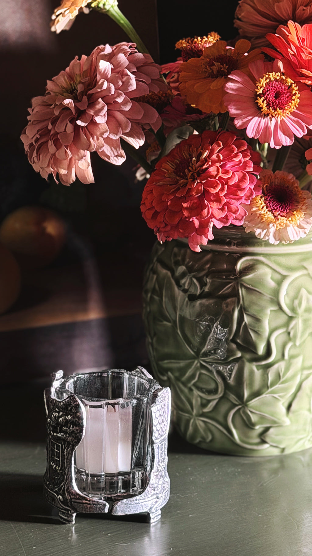 A close-up still life of a ceramic green vase filled with pink, coral, and orange flowers, alongside a small ornate silver candle holder on a dark tabletop.