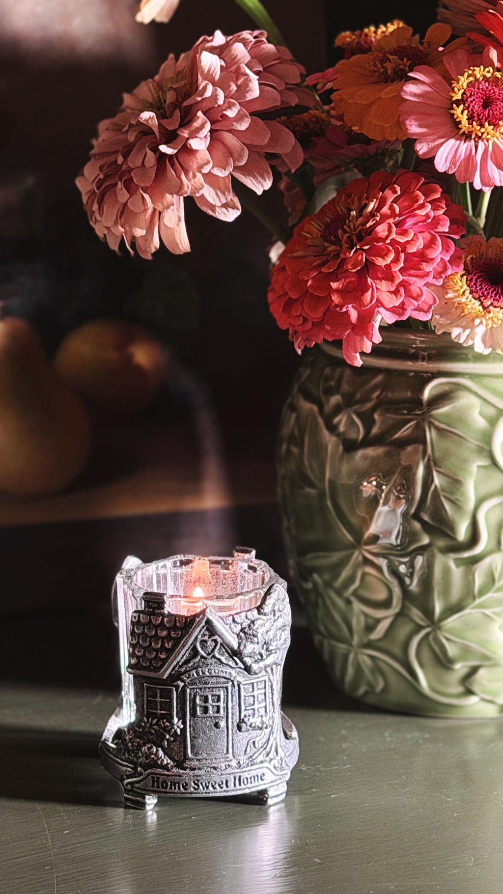A small silver house-shaped candle holder with a lit candle sits on a dark tabletop in front of a green ceramic vase filled with pink and orange flowers.
