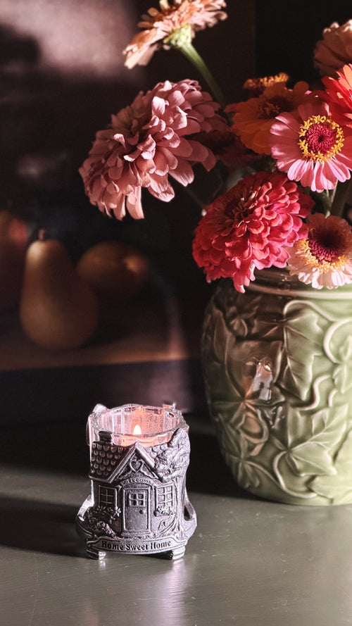 A small silver house-shaped candle holder with a lit candle sits on a tabletop in a dimly lit interior setting, with a green floral vase of pink and orange flowers in the background.