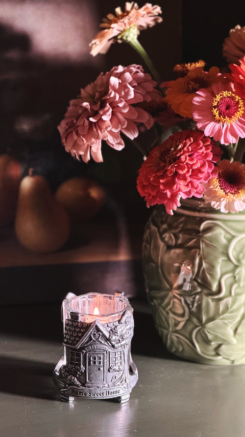 A small silver house-shaped candle holder with a lit candle sits on a tabletop in the foreground, with a vase of pink and orange flowers blurred in the background.