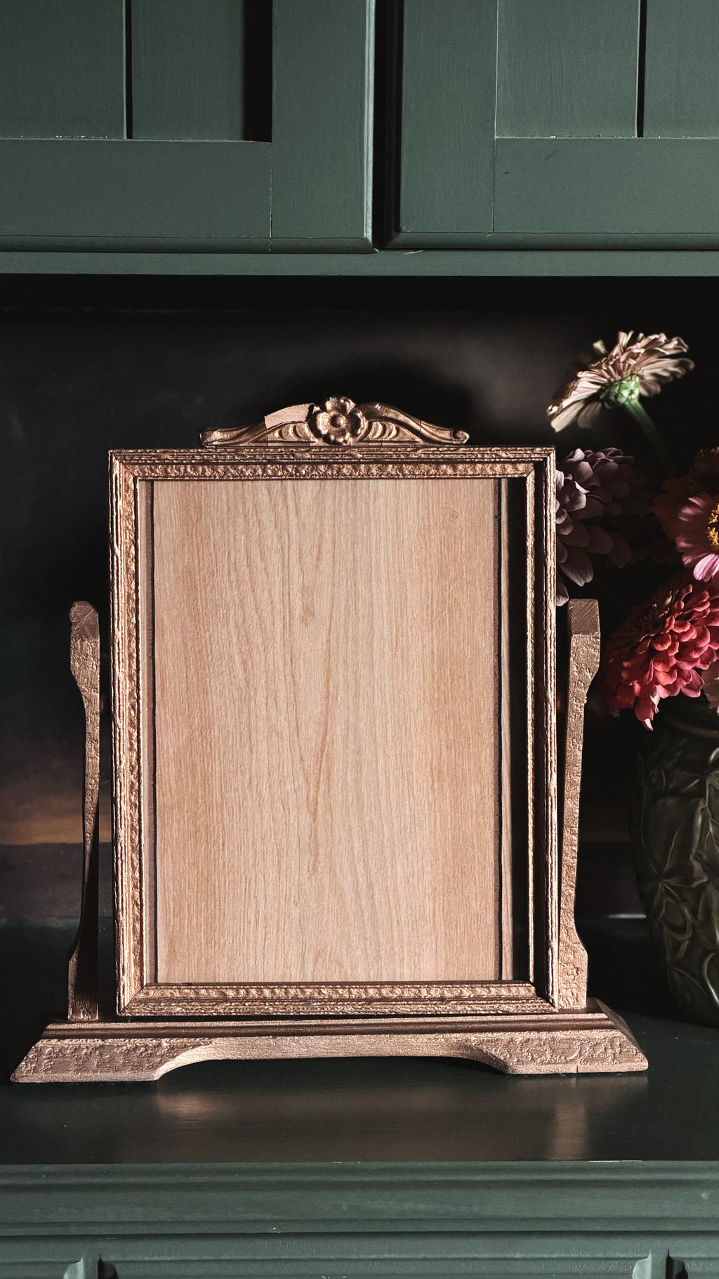 An ornate rectangular wooden picture frame with a matching stand, displayed on a dark green cabinet next to a vase of flowers.
