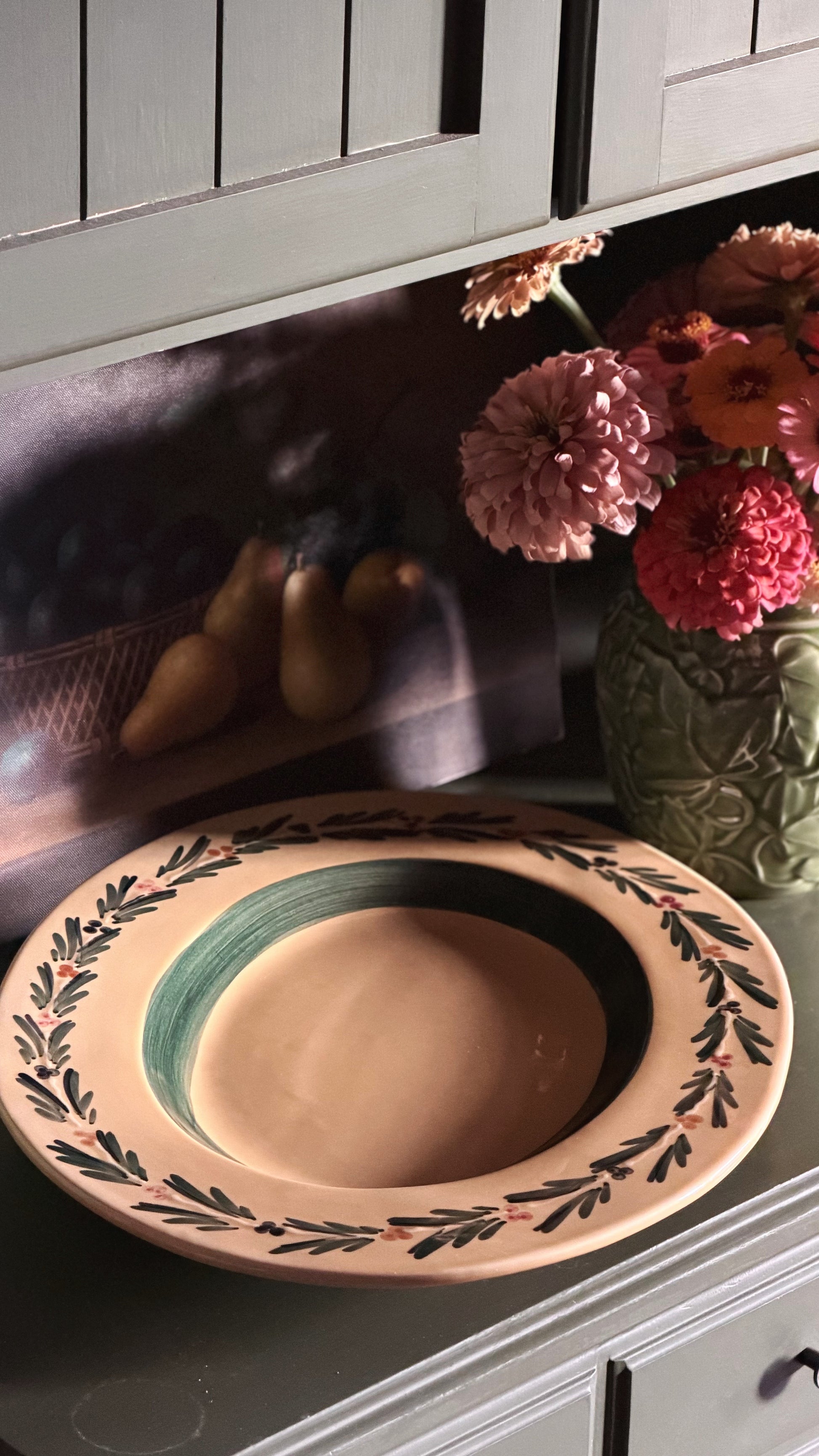 A rustic ceramic serving platter with a hand-painted floral border, displayed on a kitchen counter next to a vase of flowers.