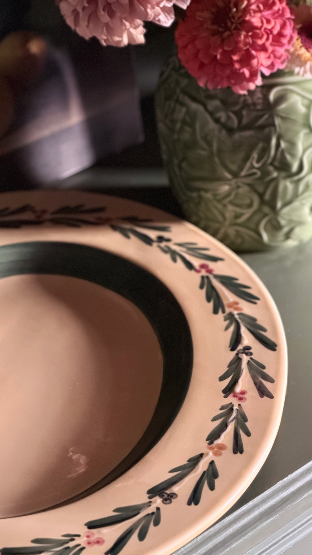 A close-up of a Vintage Williams Sonoma Italian Serving Platter with a dark green rim, and a hand-painted border of green leaves and small pink flowers. A green textured vase with pink flowers sits in the background on a tabletop.