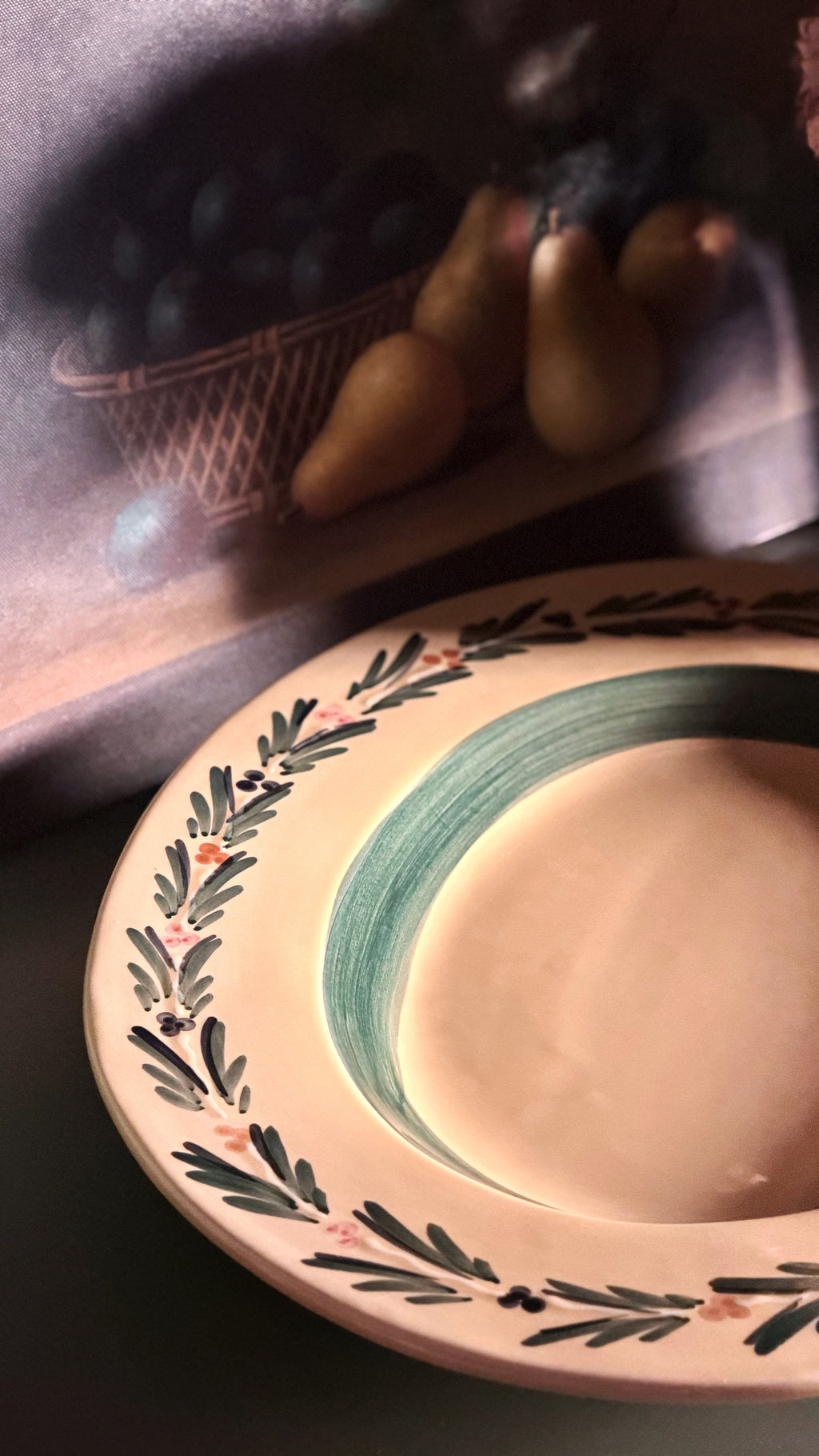 A close-up of a Vintage Williams Sonoma Italian Serving Platter with a dark green painted band, and a leafy border with small pink floral accents, set against a dark background with a blurred still life of pears and grapes.