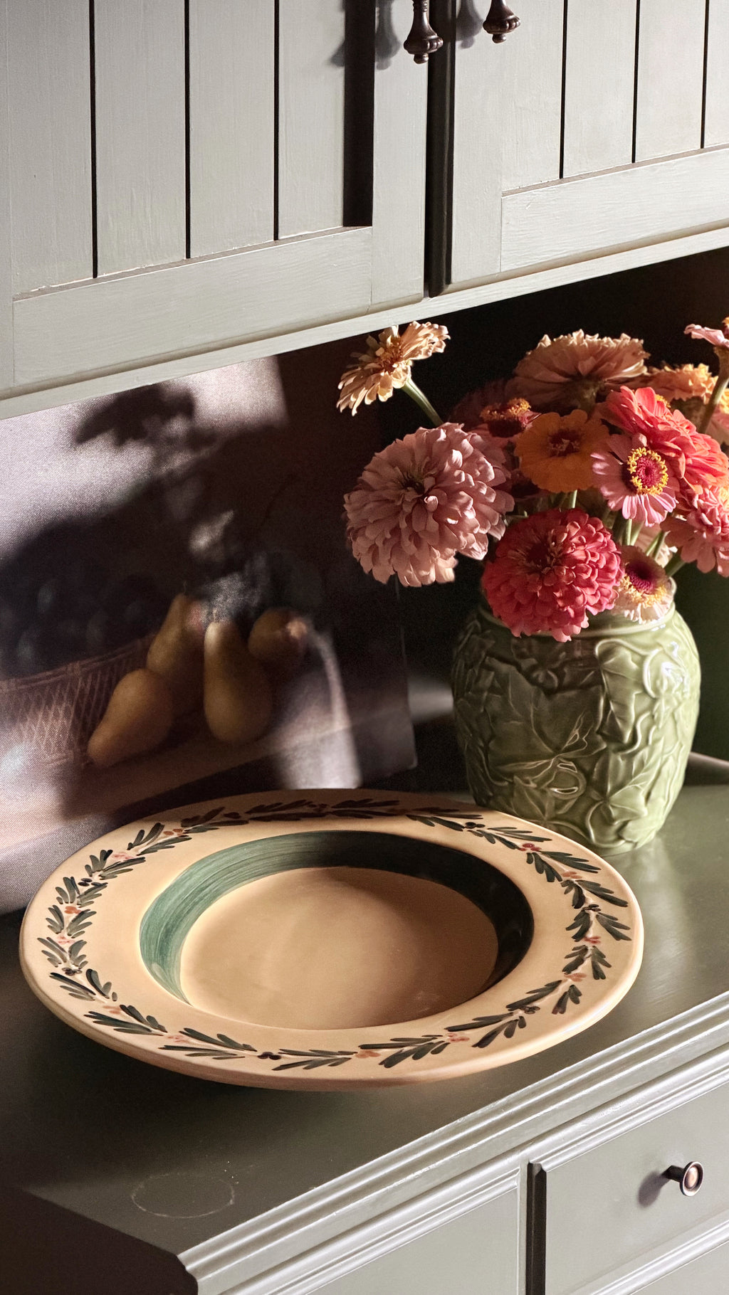 A Vintage Williams Sonoma Italian Serving Platter with a dark green inner band, and a leafy border, displayed on a kitchen counter next to a green floral vase with pink and orange flowers.